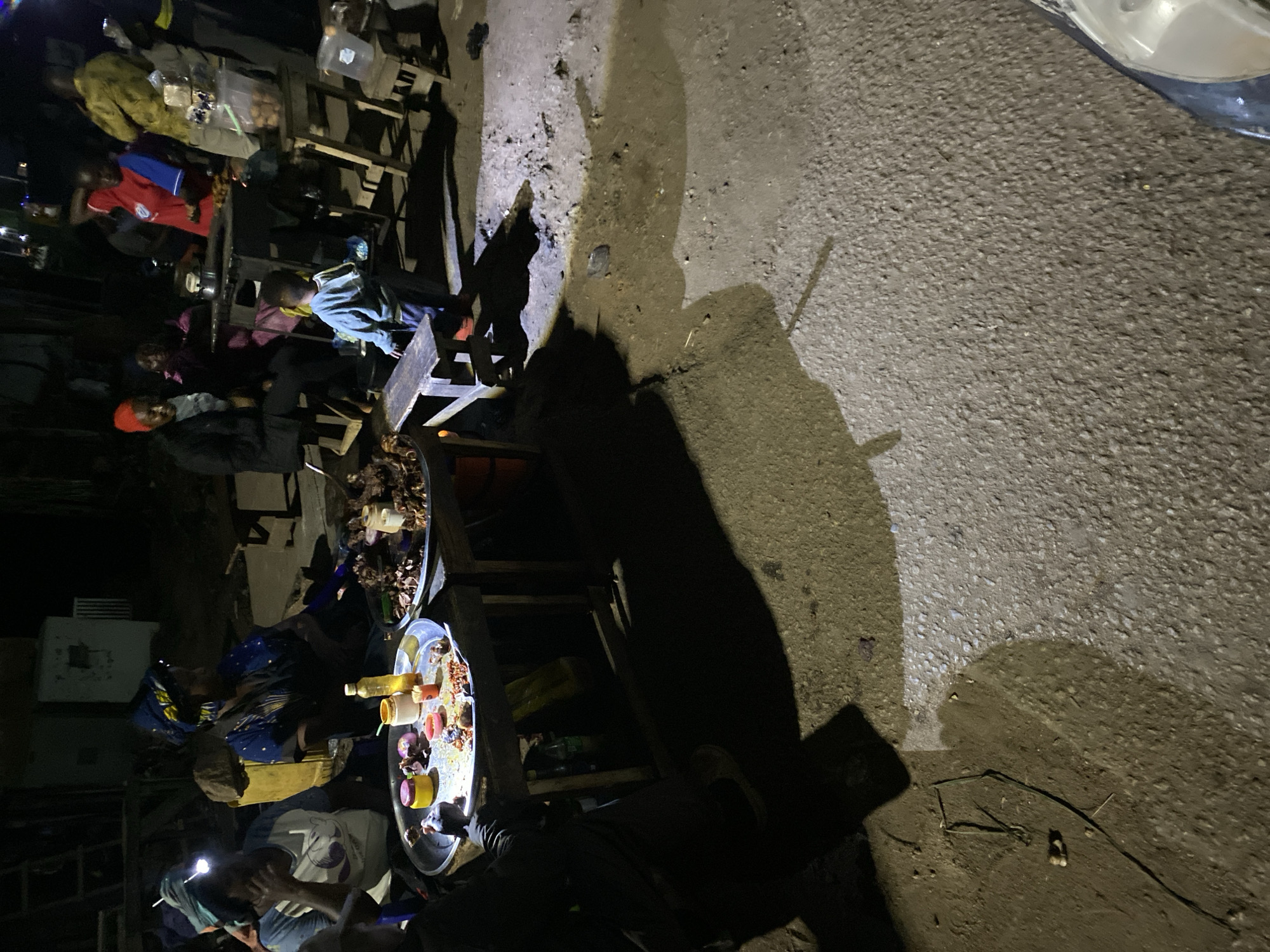 The image depicts a street scene at night in Bukuru, Nigeria. Several people are gathered around food vendors, who are selling food from tables illuminated by individual lights. One vendor is wearing a headlamp. The food appears to be arranged on trays. The street is paved, with visible shadows cast by the tables and people. A portion of a vehicle, potentially a car, is visible in the upper right corner. The environment appears to be dimly lit.