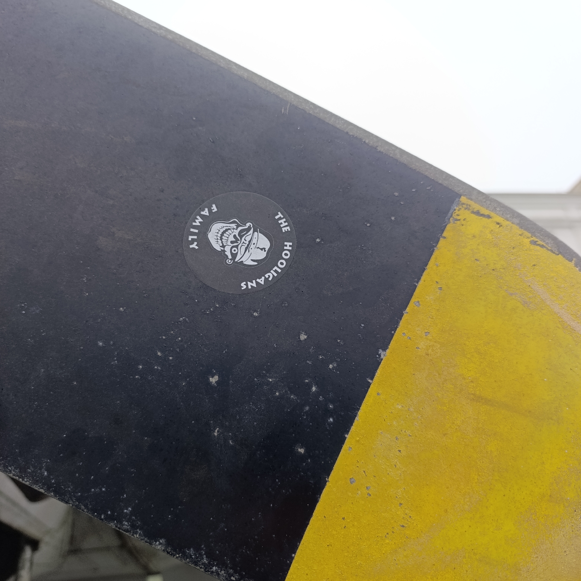 The image captures a close-up view of a weathered, dark object, likely a component of heavy machinery or an aircraft, set against a bright, indistinct background, possibly an overcast sky. The object's surface is a textured, matte black, showing signs of wear, dirt, and light-colored specks. A prominent diagonal section on the right side of the object is painted in a faded, chipped yellow, indicating age and exposure.
A circular, semi-transparent sticker is affixed to the black portion of the object. The sticker features white graphics and text. Central to the design is a stylized skull, depicted wearing a helmet or protective headgear, possibly with a ram's horn or similar motif extending from its side. Arcing above the skull on the left is the word "FAMILY," while the words "THE HOOLIGANS" are curved along the right side, reading downwards.
The scene is static, with no visible activity or people. The overall appearance suggests an outdoor or industrial environment. The location context for this media file is Verkhnyaya Pyshma, Russia.