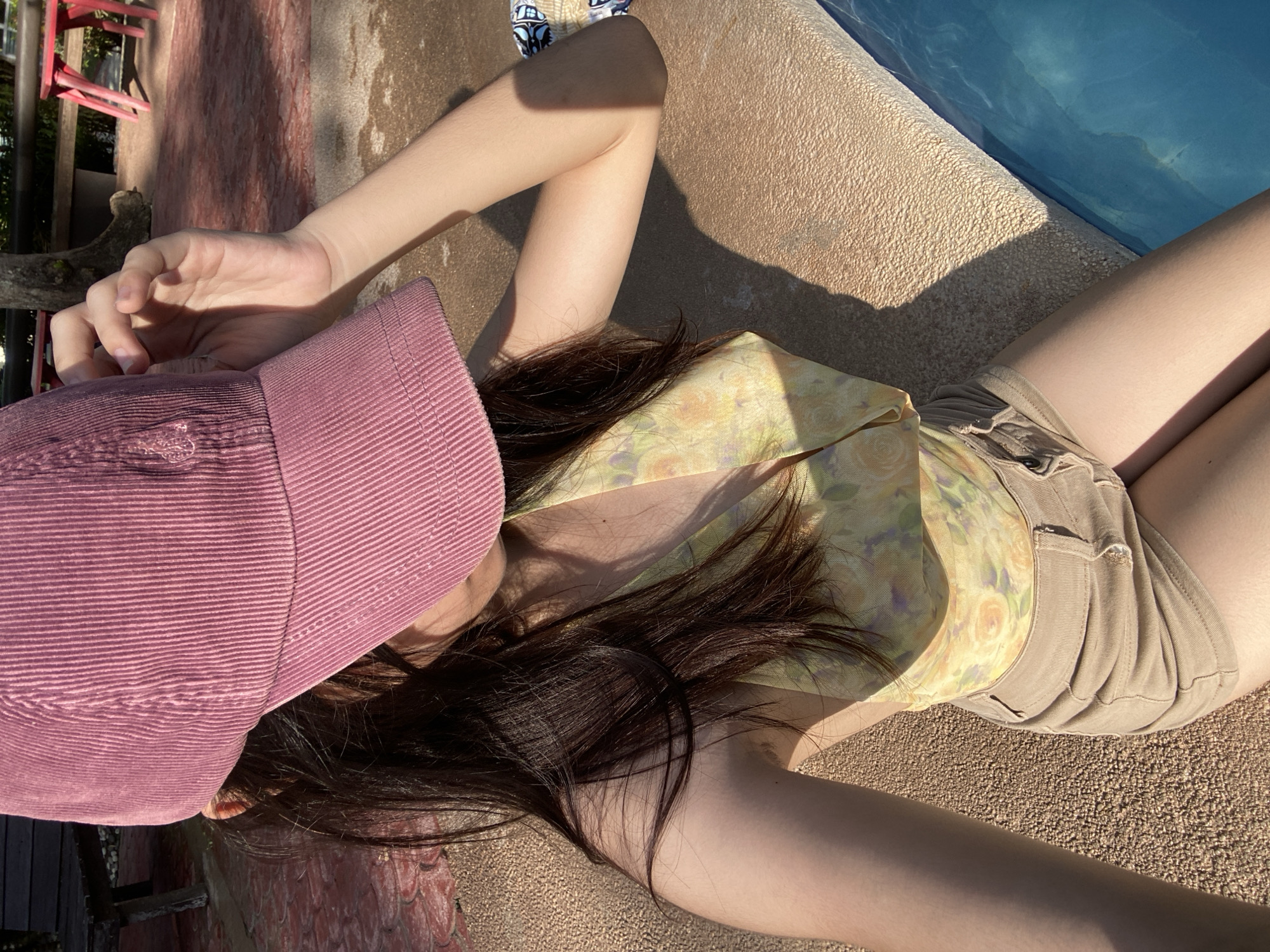 A person lies near a swimming pool. The person is wearing a pink cap, a yellow floral top, and khaki shorts. The location is likely outdoors, possibly in or near Banaybanay, Philippines given the prompt.