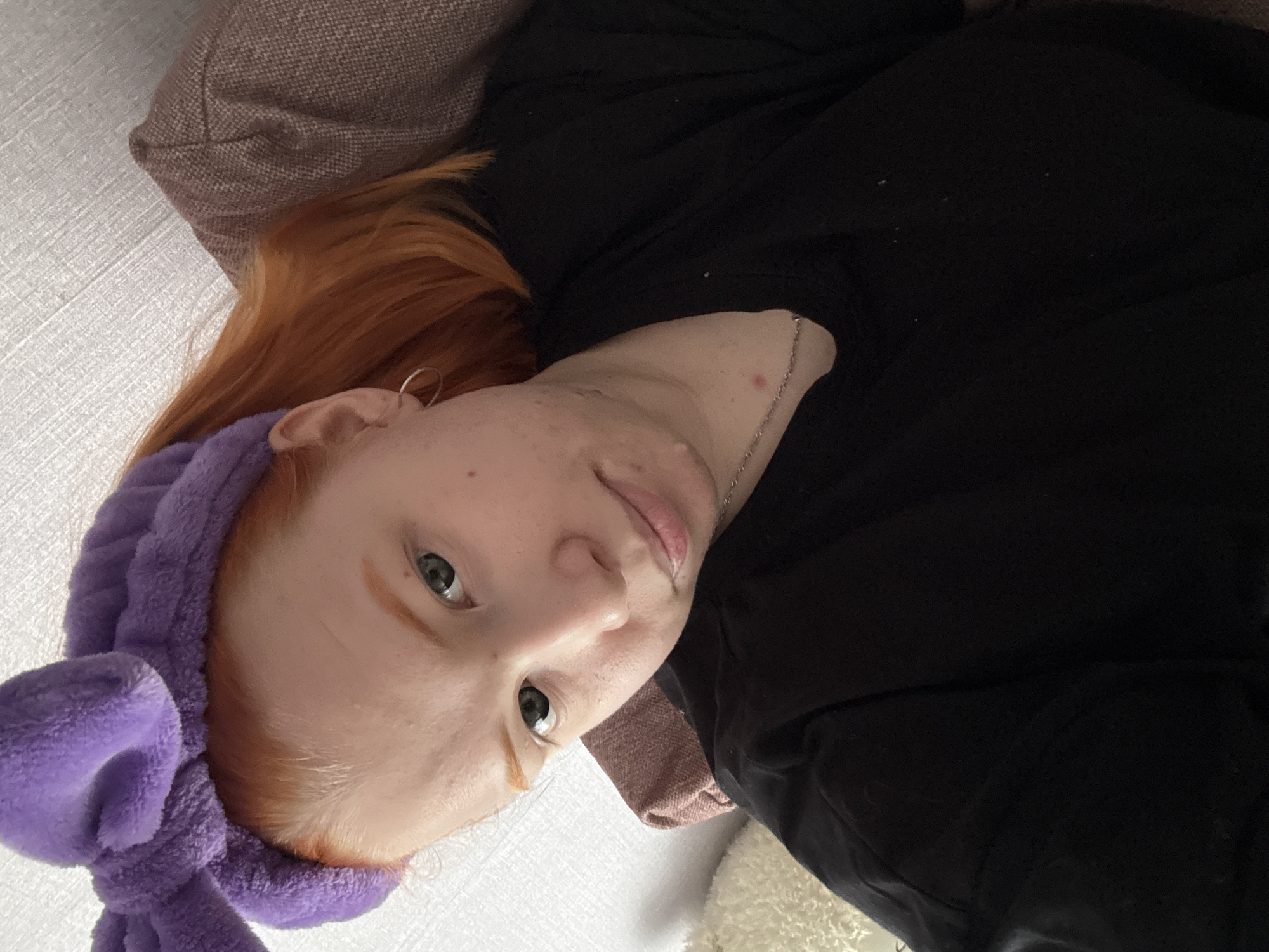 A young person with red hair and light skin is lying on a white surface with a brown pillow. They are wearing a purple headband with a bow. The person is wearing a black shirt. The location is unknown.