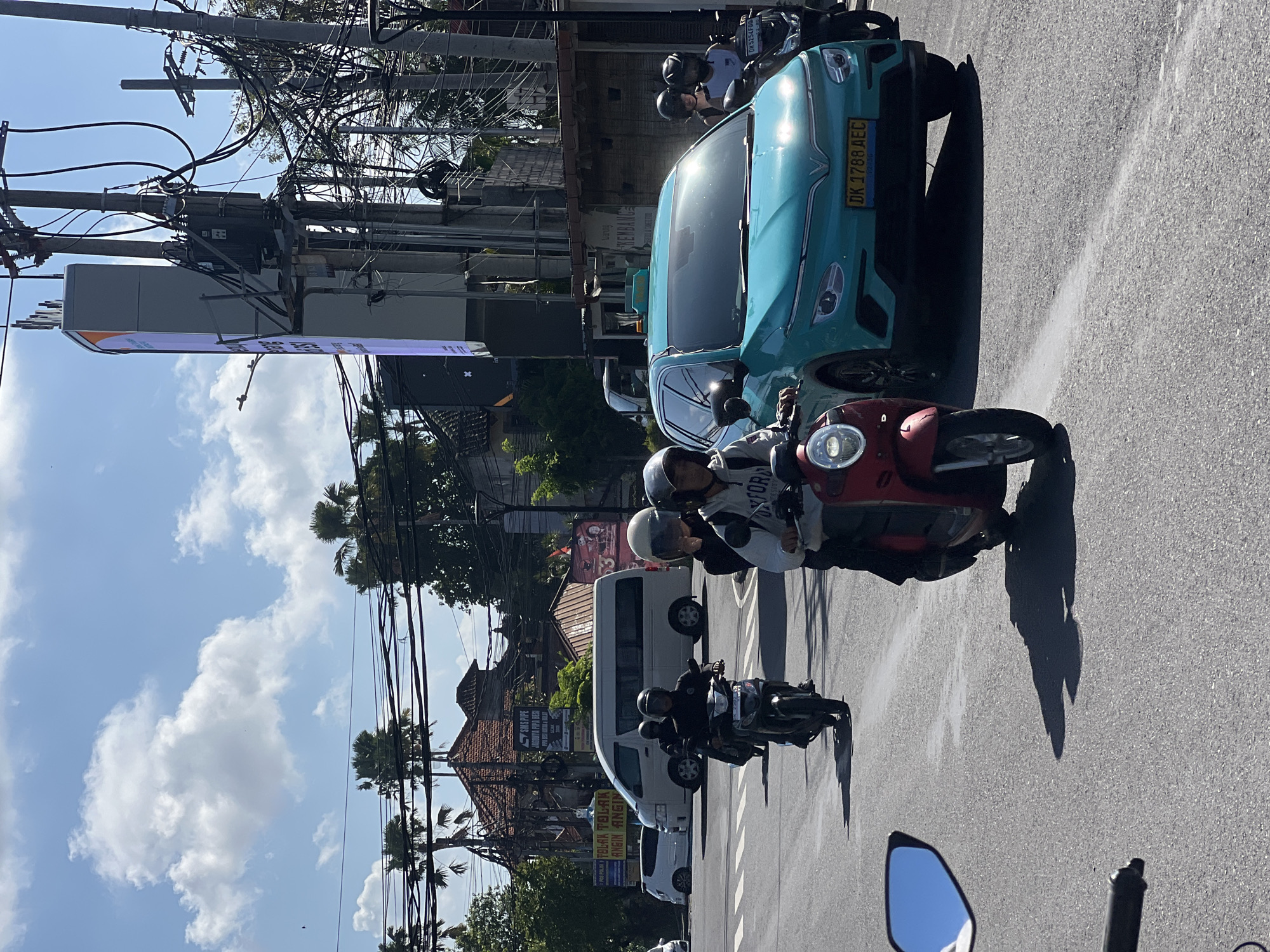 The image depicts a busy street scene in Denpasar, Indonesia, likely during daytime as indicated by the bright sunlight and shadows. The foreground shows the handlebars and a wing mirror of a motorcycle, suggesting the photograph is taken from a moving vehicle. Several other vehicles are visible, including a bright teal car with a yellow license plate and a white van. Multiple individuals are riding motorcycles, most wearing helmets. One person on a red scooter is prominent in the middle ground. Palm trees and traditional Indonesian architecture, including red-tiled roofs, are present in the background, interspersed with utility poles and electrical wires, characteristic of urban environments in Indonesia. The sky is blue with scattered white clouds.