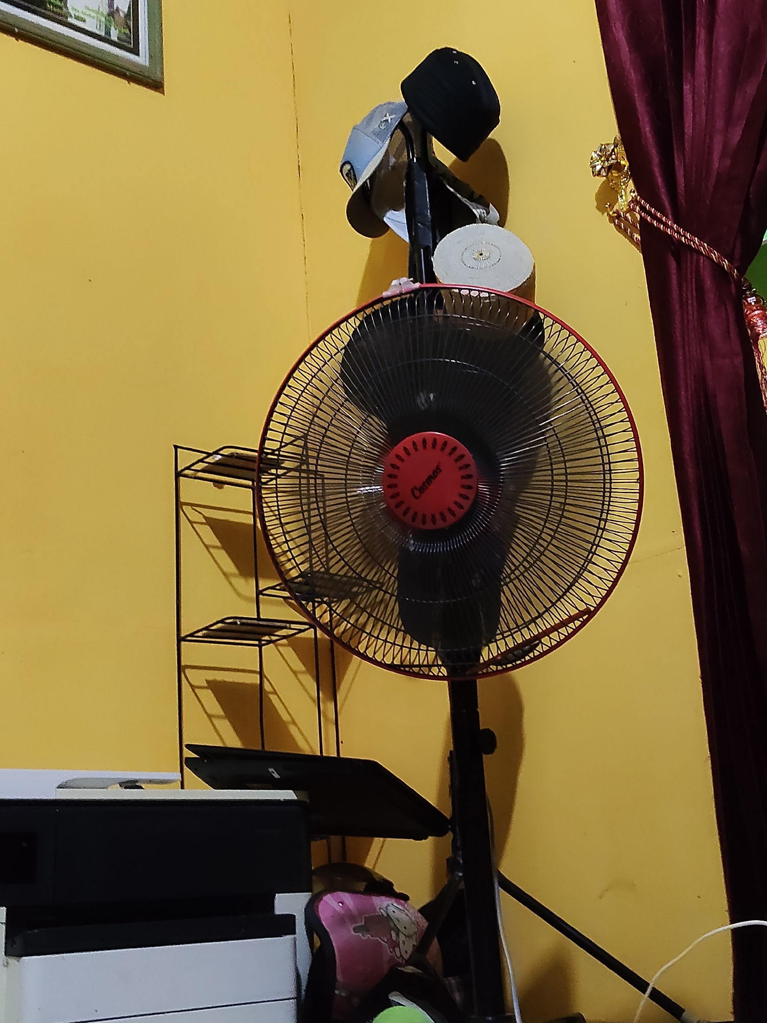 In a room with yellow walls, a black oscillating fan with a red central hub stands on a black pole. On top of the fan's pole are several caps, including a black baseball cap and a grey cap. To the left of the fan, a black metal shelf unit with three shelves is visible. In the foreground to the left, a white and black printer-like object is partially visible. To the right of the fan, a dark red curtain is draped. The scene appears to be indoors, likely in a residential setting in Tanah Grogot, Indonesia.