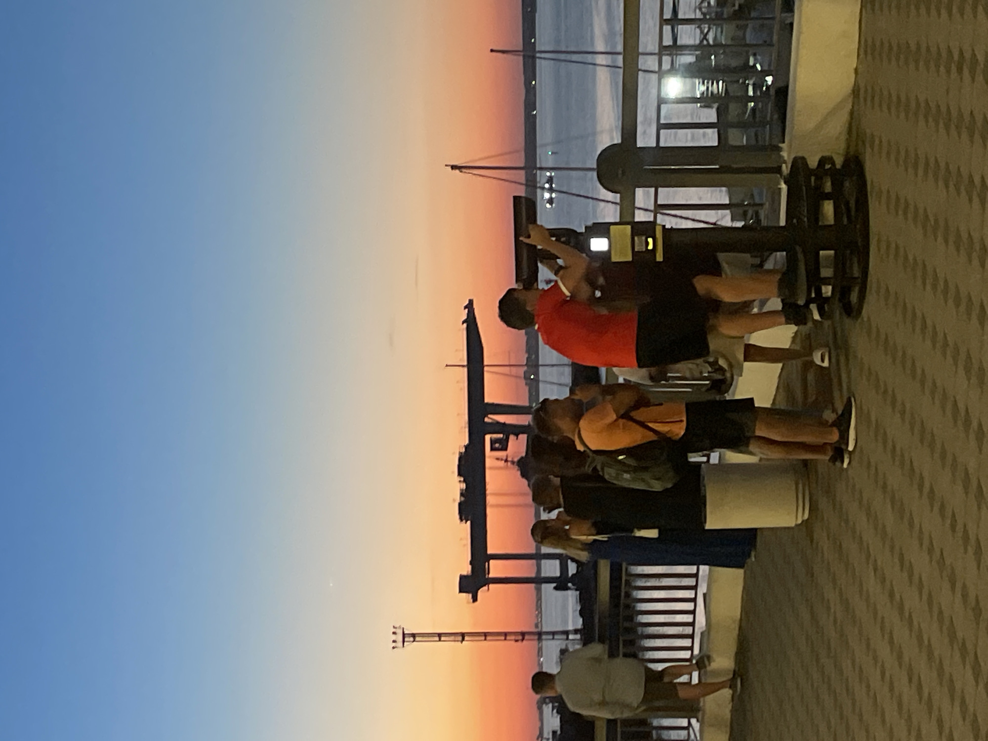 This is a vertical, medium shot of people at a waterfront during a sunset in Unknown, Unknown. In the foreground, a patterned brick walkway is visible. A black metal railing runs along the edge of the walkway, separating it from the water beyond. Several people are standing on the walkway, looking out at the water.
On the left, a person in a light-colored shirt and shorts stands with their back to the camera. To their right, a woman in a blue dress and two people in dark clothing stand together, with one of them holding a backpack. Further to the right, a person in an orange t-shirt and black shorts is looking through a pair of binoculars mounted on a stand. Another person in a red shirt and black shorts stands next to them.
In the background, a body of water is visible, with several sailboats docked or moored. A large industrial crane is also prominent in the background, silhouetted against the colorful sunset sky. The sky transitions from a pale blue at the top to vibrant shades of orange and pink near the horizon.
