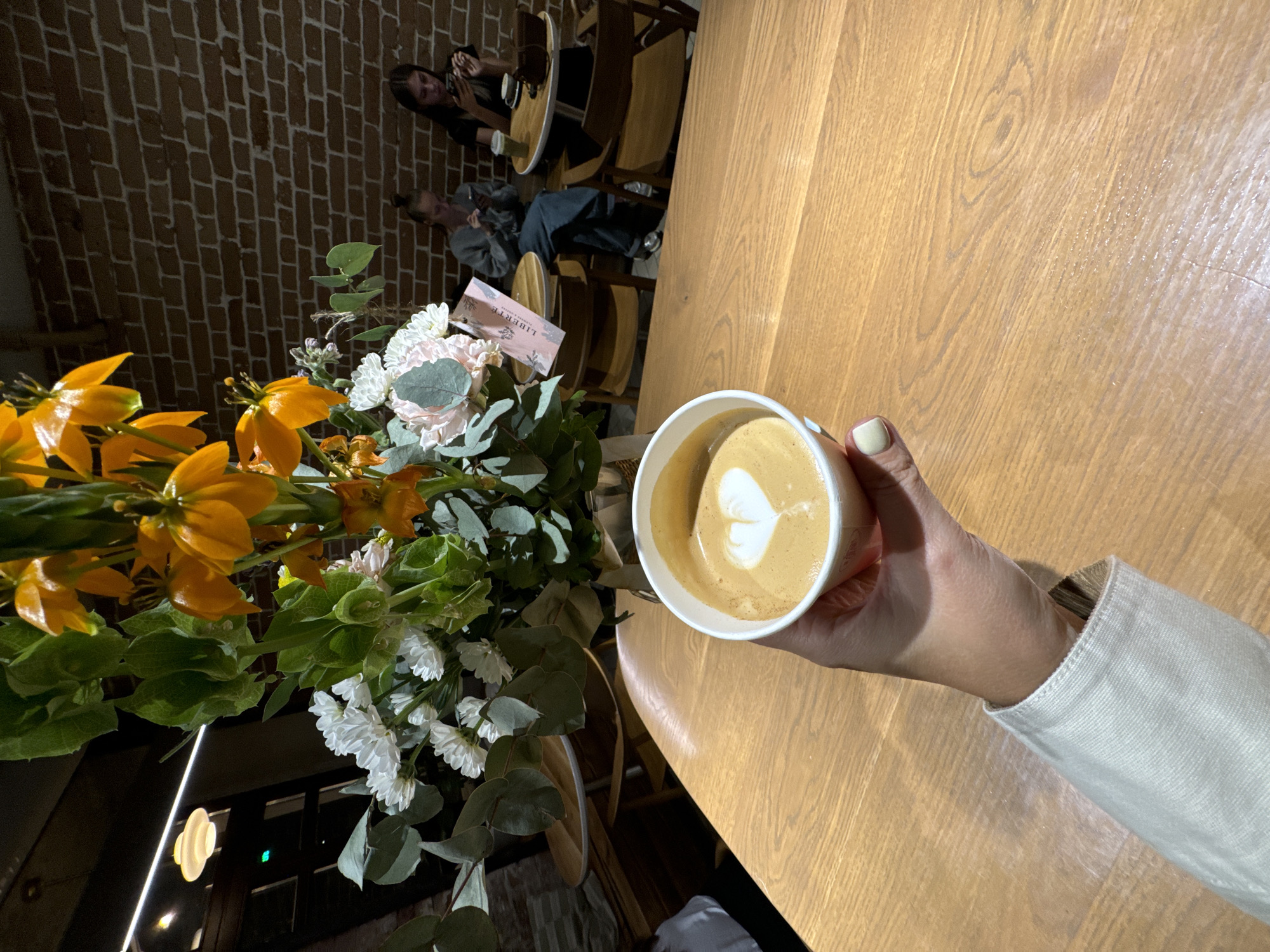 A person's hand in a light-colored jacket sleeve holds a paper cup of coffee with latte art shaped like a heart. The cup is on a wooden table in what appears to be a cafe. In the background, there is a bouquet of flowers, including orange and white blooms, and two young women sitting at tables, looking at their phones. The wall behind them is made of brick. The cafe setting is consistent with establishments found in urban areas, such as the city of Krasnoyarsk, Russia.