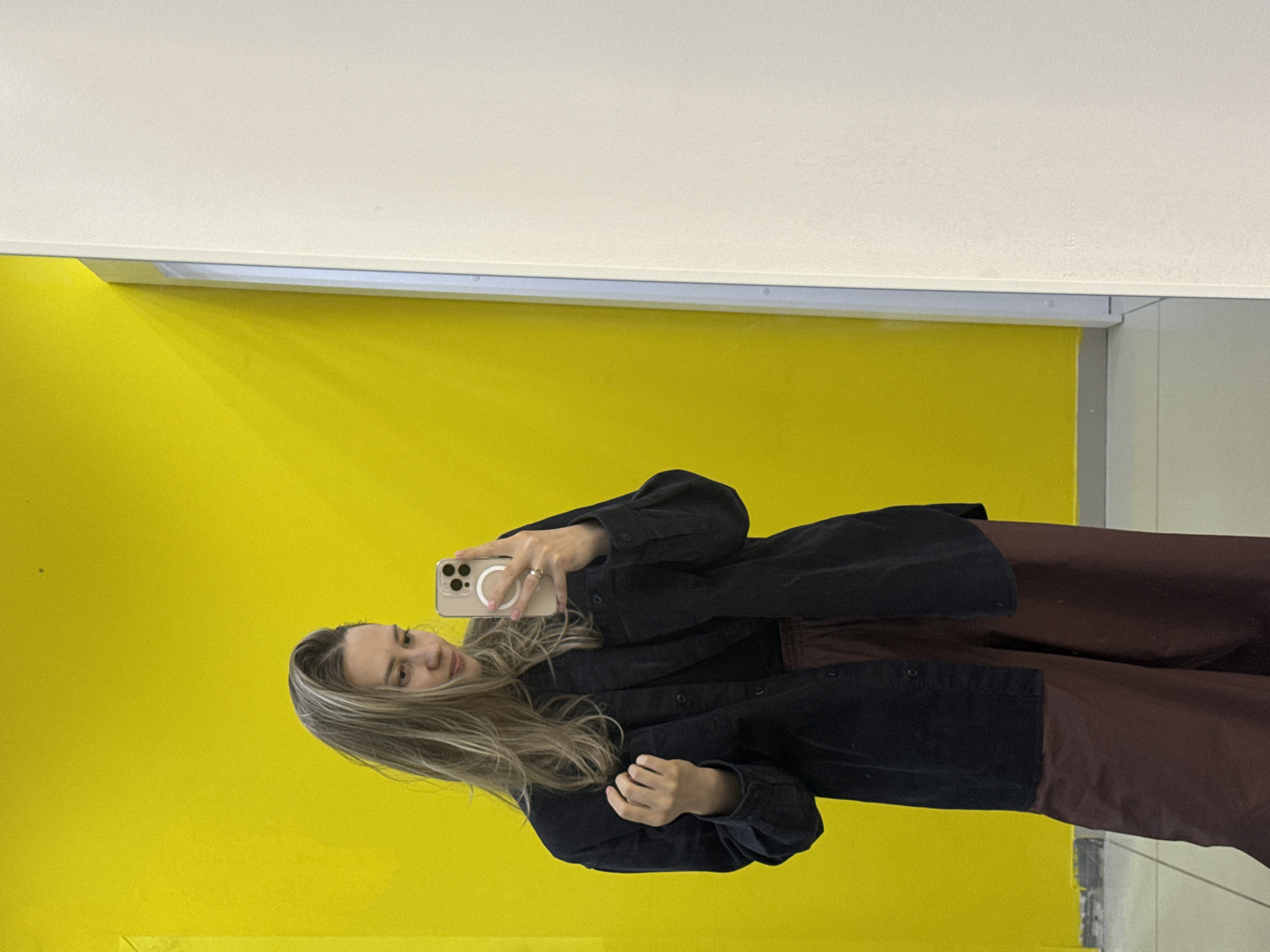 This image is a mirror selfie featuring a young woman with long, light brown hair in an indoor setting. She is wearing a dark, likely black or navy, long-sleeved button-down shirt or jacket, layered over what appears to be a dark reddish-brown top or dress. Her right hand, adorned with a ring on her ring finger, holds a light-colored smartphone (likely an iPhone given its design and MagSafe ring) up to capture the photograph. Her left hand rests at her side.
The woman is looking towards the phone with a soft, relaxed expression, possibly winking or blinking with her left eye.
The background primarily consists of a vibrant, bright yellow wall, occupying most of the space behind her. Above the yellow wall is a plain white ceiling, separated by a crisp white horizontal trim or light fixture. On the far right, a silver or grey vertical frame is partially visible, reinforcing the context of a mirror or glass partition. The lighting within the scene is bright and even, typical of an indoor public or commercial environment. The overall setting appears modern and clean, located within Chaikovsky, Russia. No visible text is present in the image.