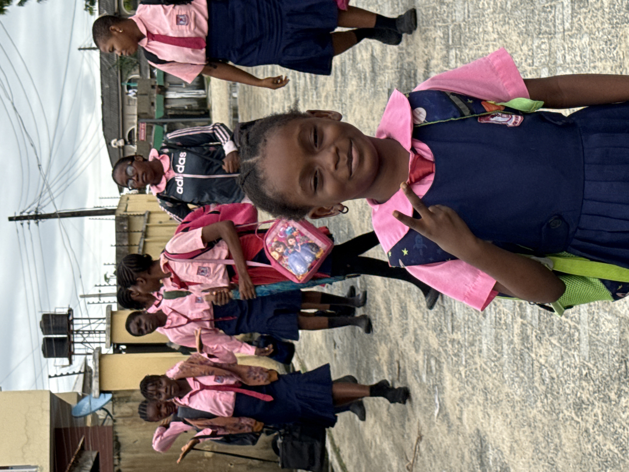 A line of young girls wearing school uniforms are standing on a paved surface in Ado, Nigeria. The uniforms consist of a light pink shirt and a navy blue skirt. The girl in the foreground is giving a peace sign and smiling at the camera. The background shows buildings and objects like water tanks. Some of the girls are carrying backpacks. One individual is wearing an Adidas tracksuit. The sky is overcast.