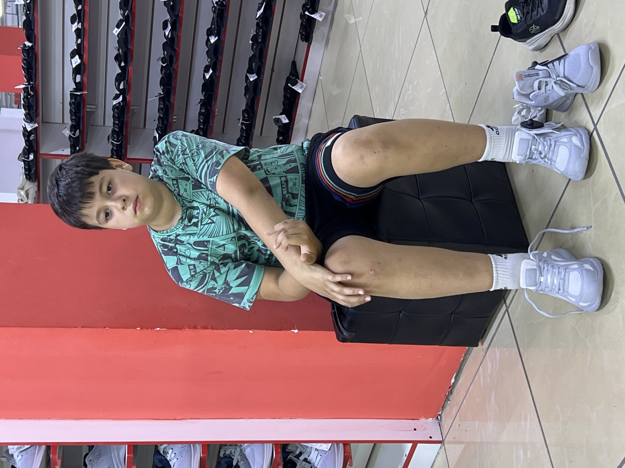 The image depicts a young boy sitting on a black cube in what appears to be a shoe store, possibly located in Krasnodar, Russia. The boy is wearing a teal t-shirt with a graphic print and black shorts with a colorful striped waistband. He is barefoot except for white socks. In front of him are several pairs of white sneakers, suggesting he is trying them on.
The background reveals shelves filled with footwear, primarily black sandals, indicating a wide selection of shoes. A bright red wall is prominent to the left of the boy, and a white shelf unit with more shoes is visible further left. The floor is tiled with a light-colored, reflective material. The overall lighting suggests an indoor commercial setting.