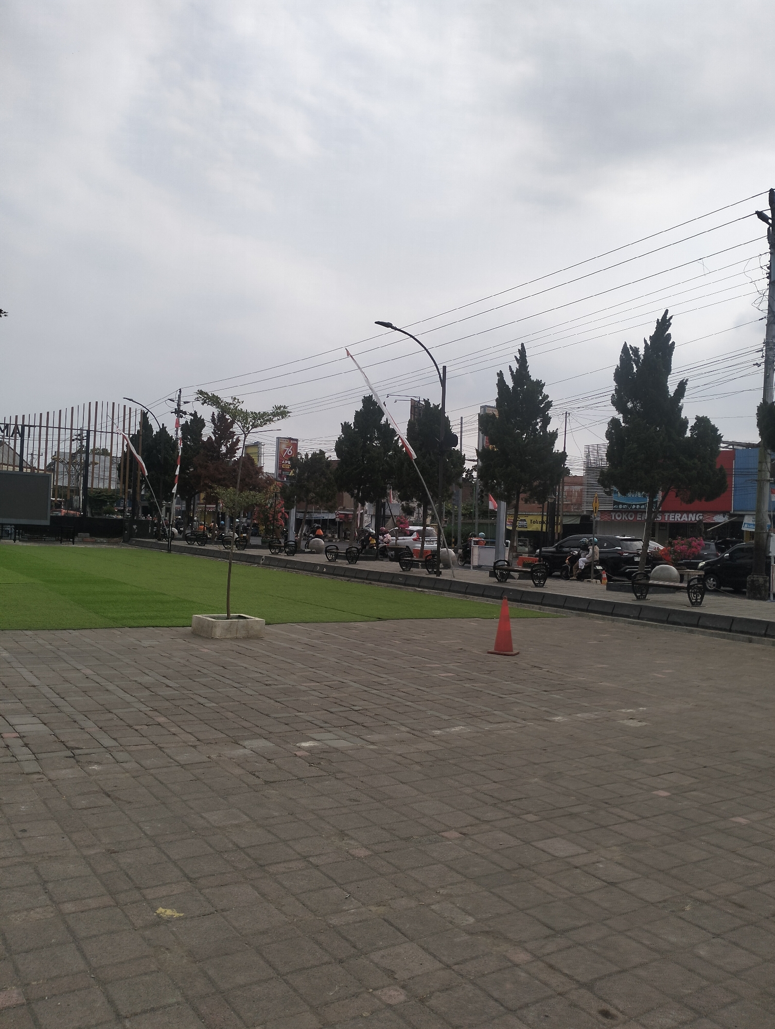 The image depicts a paved public square in Purbalingga, Indonesia, adjacent to a street with vehicles and trees. In the foreground, a paved area of grey square tiles leads to a well-maintained patch of bright green artificial turf. A young sapling in a concrete planter sits on the turf. To the left, a large black screen is visible behind a metal fence.
Along the street, there are several trees with dark green foliage, characteristic of cypress trees, lining the area. A traffic light pole with multiple wires stretches across the upper portion of the scene. Several motorcycles and cars are parked or moving along the street. Some individuals are visible, primarily near the parked vehicles. A red traffic cone is placed on the tiled ground.
In the background, buildings with various signage and architectural styles are present. One building prominently displays a "76 APK 2023" sign, possibly related to a local event or election. Indonesian flags are also visible, with red and white stripes on poles. The sky is overcast with a pale grey, cloudy appearance. The overall scene suggests a public space in a moderately busy urban environment in Purbalingga.