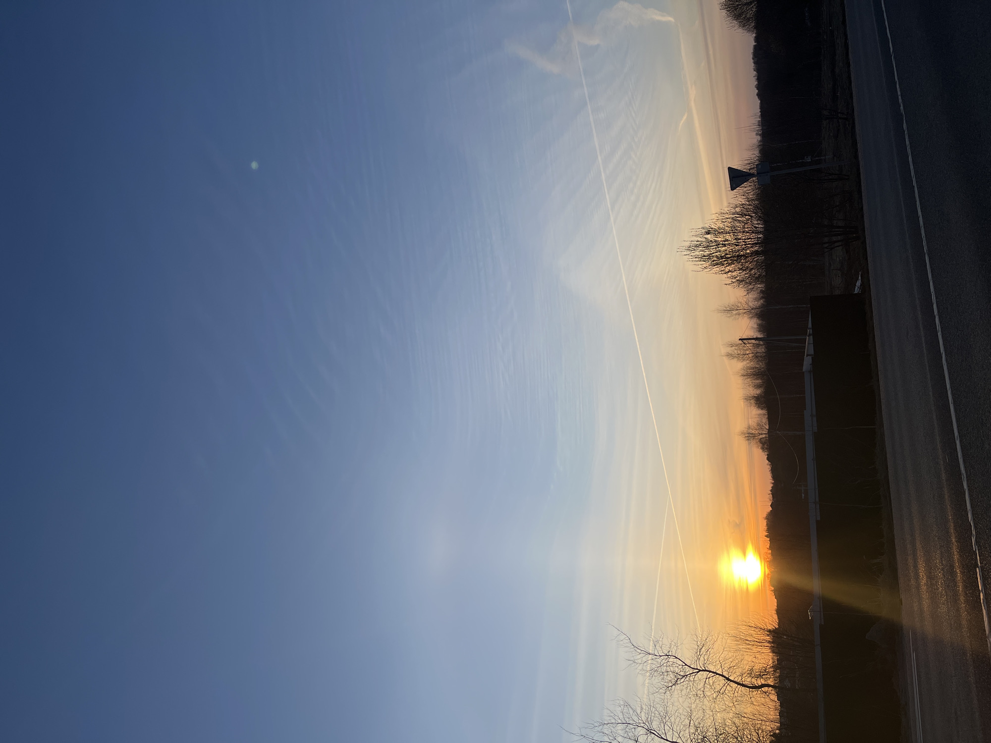 The image shows a landscape with the sun low on the horizon, appearing to be either a sunrise or sunset. The sky transitions from a deep blue in the upper left to orange and yellow near the horizon. Thin contrails from airplanes are visible in the sky. A dark treeline silhouettes against the bright horizon, along with a few bare trees in the foreground. A paved road runs along the right edge of the image. Based on the landscape and presence of a road, it's an outdoors scene possibly near the city of Zaovrazh'e, Russia, but lacking more details, it is not possible to know more.