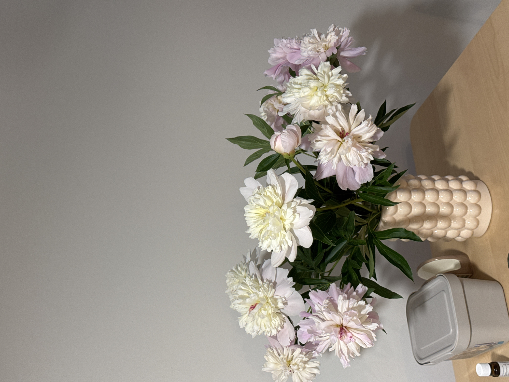 A bouquet of light pink and white peonies is arranged in a textured, light-colored vase. The peonies are in various stages of bloom, with some fully open and others still budding. The vase sits on a light brown surface, possibly a table, next to a light grey square tin with a lid. A small glass bottle with a dark cap and visible liquid, likely an essential oil or medicine, is also on the surface. The background is a plain, light grey wall, creating a simple and elegant setting. The lighting suggests it is daytime, casting subtle shadows and highlighting the delicate petals of the flowers. The overall mood is calm and serene. There is no visible text in the image.