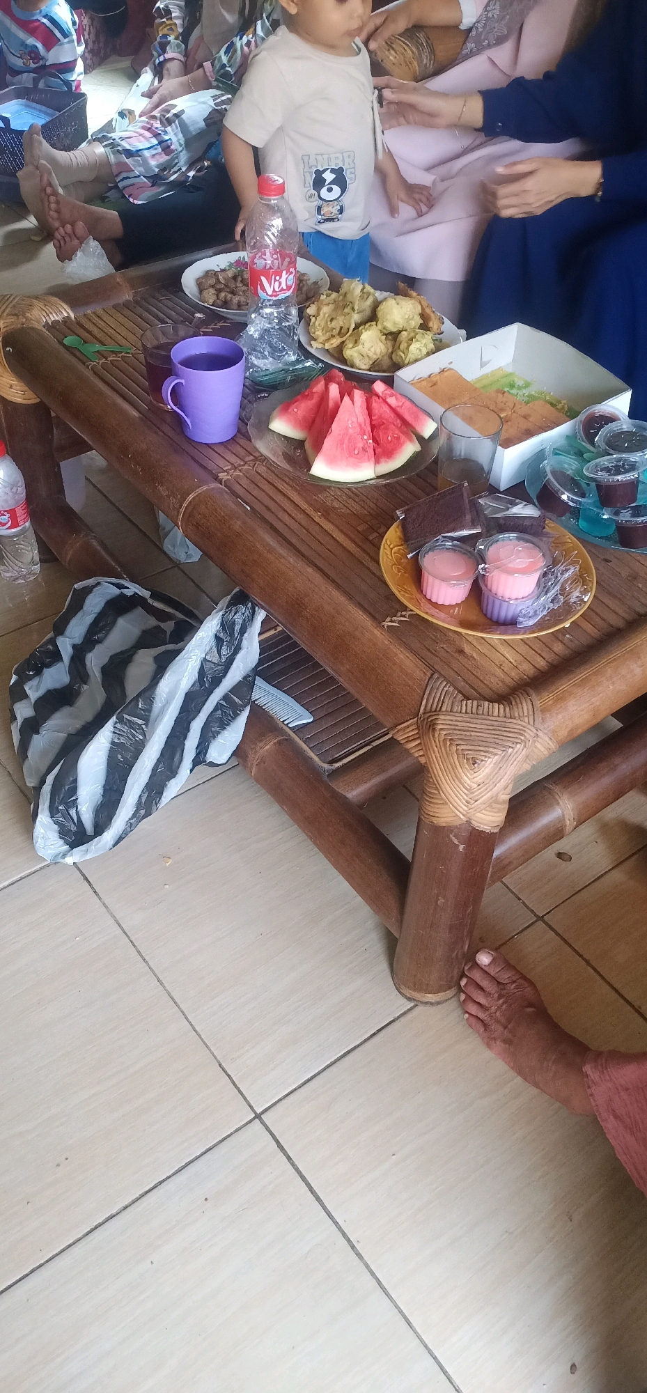 This is a close-up, eye-level, medium shot of a bamboo table laden with food and drinks, situated outdoors on a tiled floor. The scene suggests a casual gathering, possibly a meal or snack break. The foreground is dominated by the table's bamboo structure and a black and white striped plastic bag. On the table, an assortment of items are displayed: a plate of fried food, sliced watermelon on a glass plate, a box of layered cake, and several small cups containing pink desserts. A bottle of "Vita" brand water and a purple mug filled with a dark liquid are also visible. In the background, partially obscured, are glimpses of people, including a child wearing a t-shirt with a cartoon bear print, and adults dressed in traditional Indonesian attire. The lighting is natural and bright, casting soft shadows. The overall impression is one of a relaxed, informal social occasion. The image likely depicts a domestic setting or a small community gathering in Bajong, Indonesia.