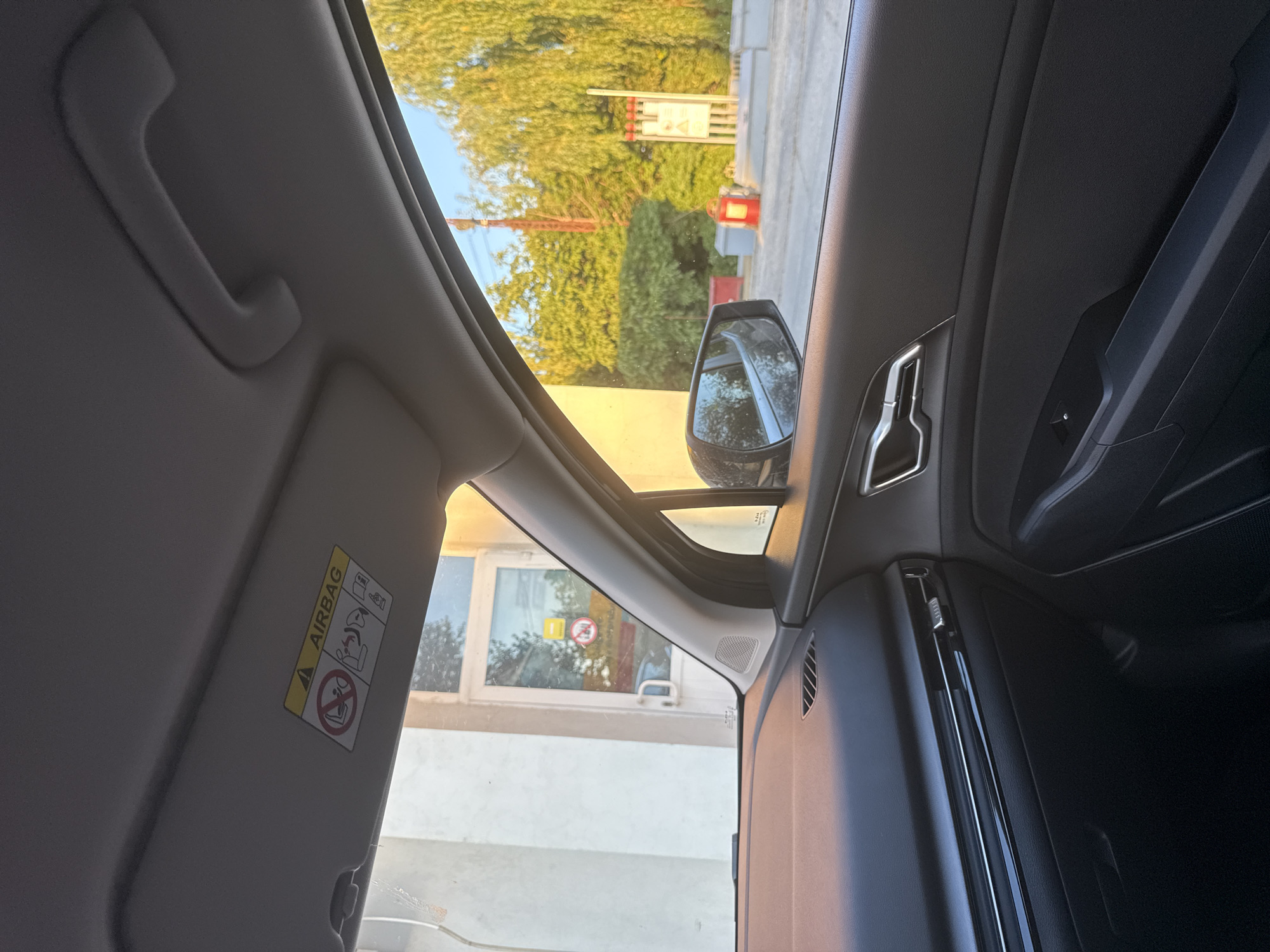 The image shows the interior of a car, looking out the passenger side window. The dashboard and door panel are visible in the foreground. Through the window, we see a building with a glass door and a yellow sign with red lettering, as well as lush green trees. A side mirror reflects a blurry image of the surroundings. A yellow sticker with warning symbols is affixed to the car's sun visor. The scene appears to be set in a city like Basarabeasca, Moldova, given the context provided. The lighting suggests it is late afternoon or early evening, with the sun casting a warm glow on the scene. There are no people visible in the image, and the car appears to be stationary.