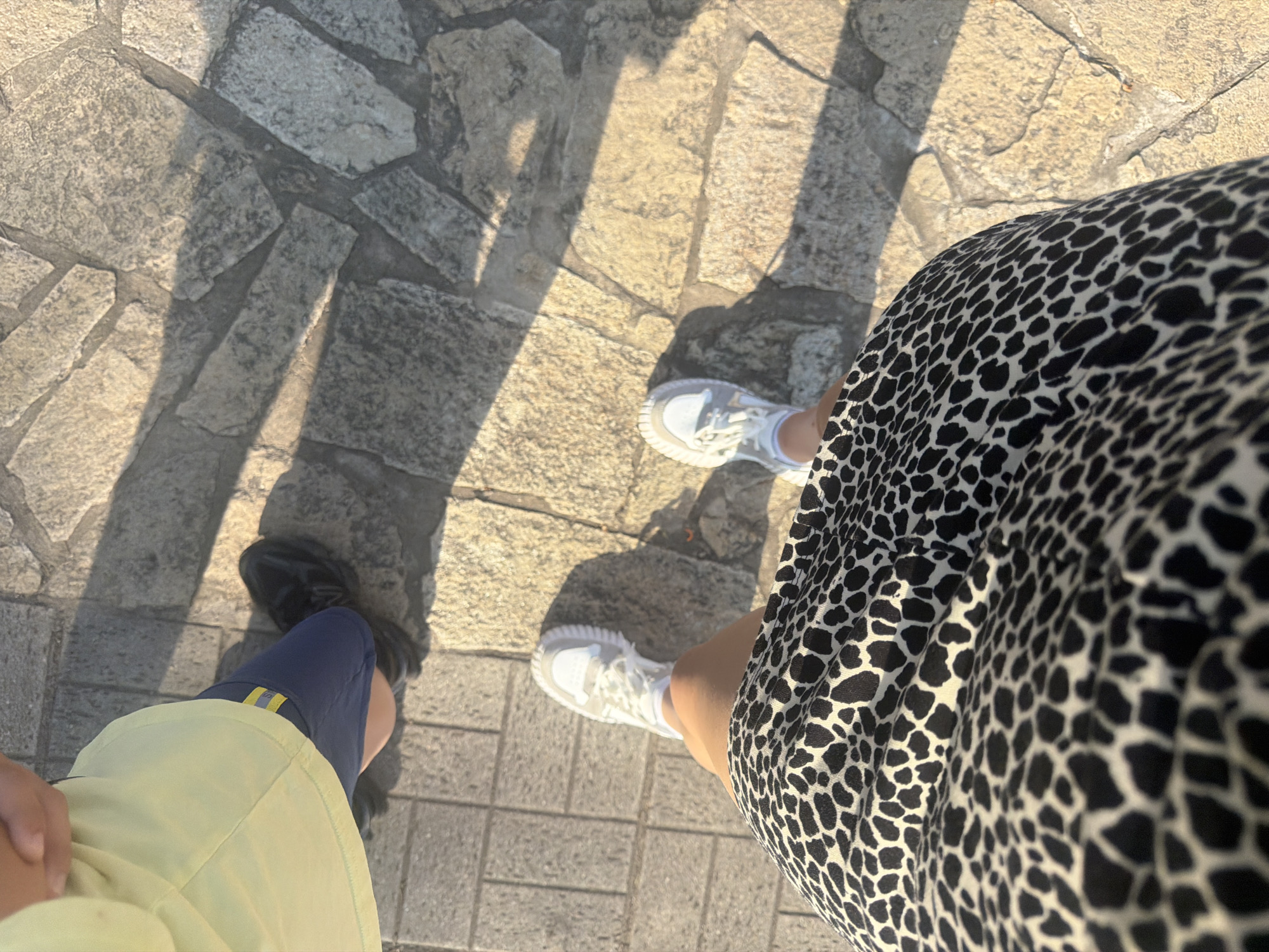 Here is a ground-level, slightly angled, outdoor shot depicting the lower portions of two individuals standing on a paved stone surface. The person on the left wears black shoes, blue shorts with a yellow stripe, and a light yellow shirt. The person on the right wears white sneakers and a black and white patterned dress. Shadows are cast across the stone surface. The location is an urban environment in an unknown city and country.