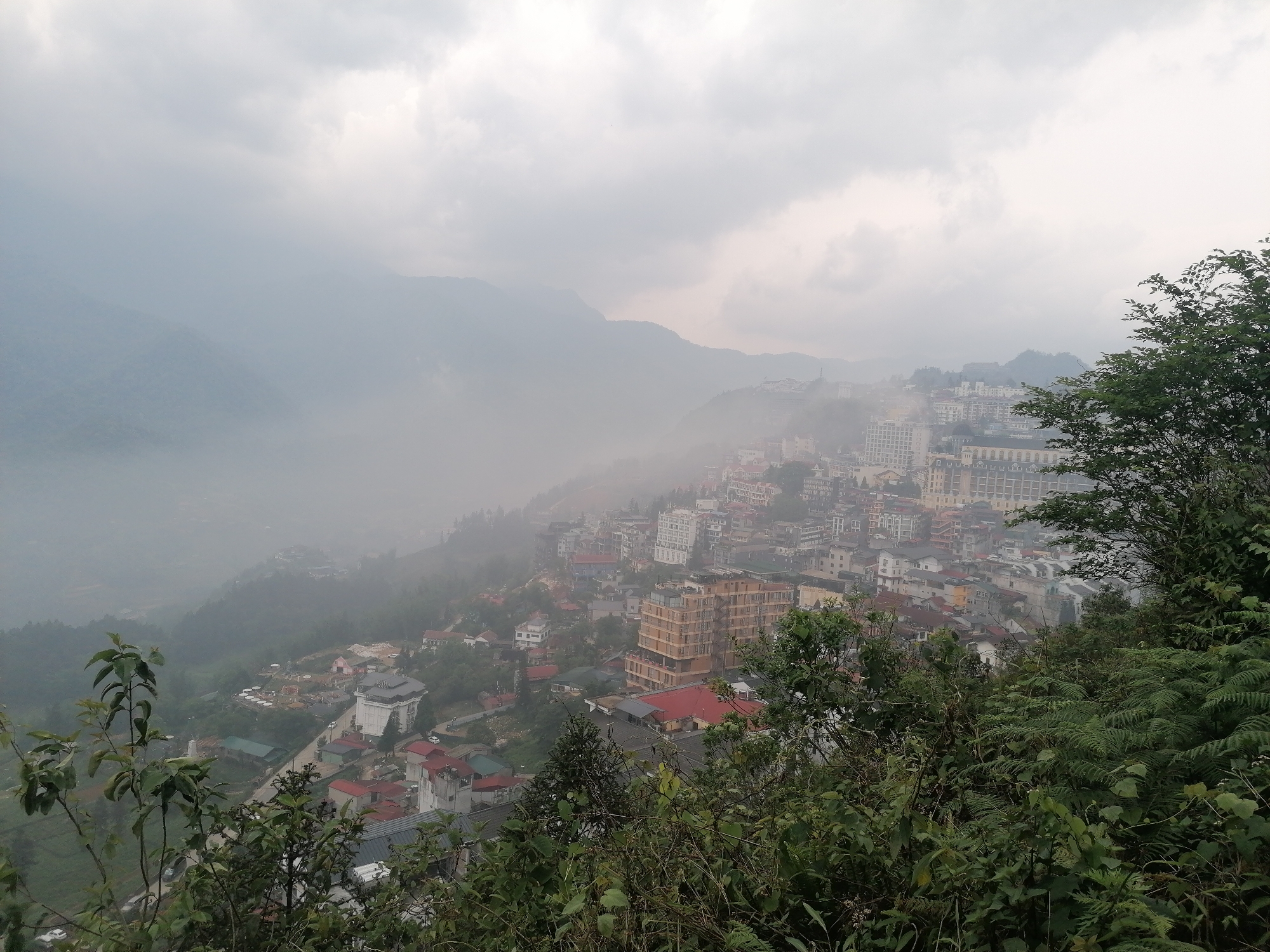 This image shows a view of Sa Pa Ward, Vietnam, from an elevated perspective, obscured by a dense fog or mist. The foreground features various green plants and foliage. The middle ground shows a cityscape with buildings of varying heights and architectural styles. The overall color palette of the city is muted with colors like white, red, and yellow visible. In the background, there are hazy mountains barely visible through the fog. The sky is overcast with grey clouds.