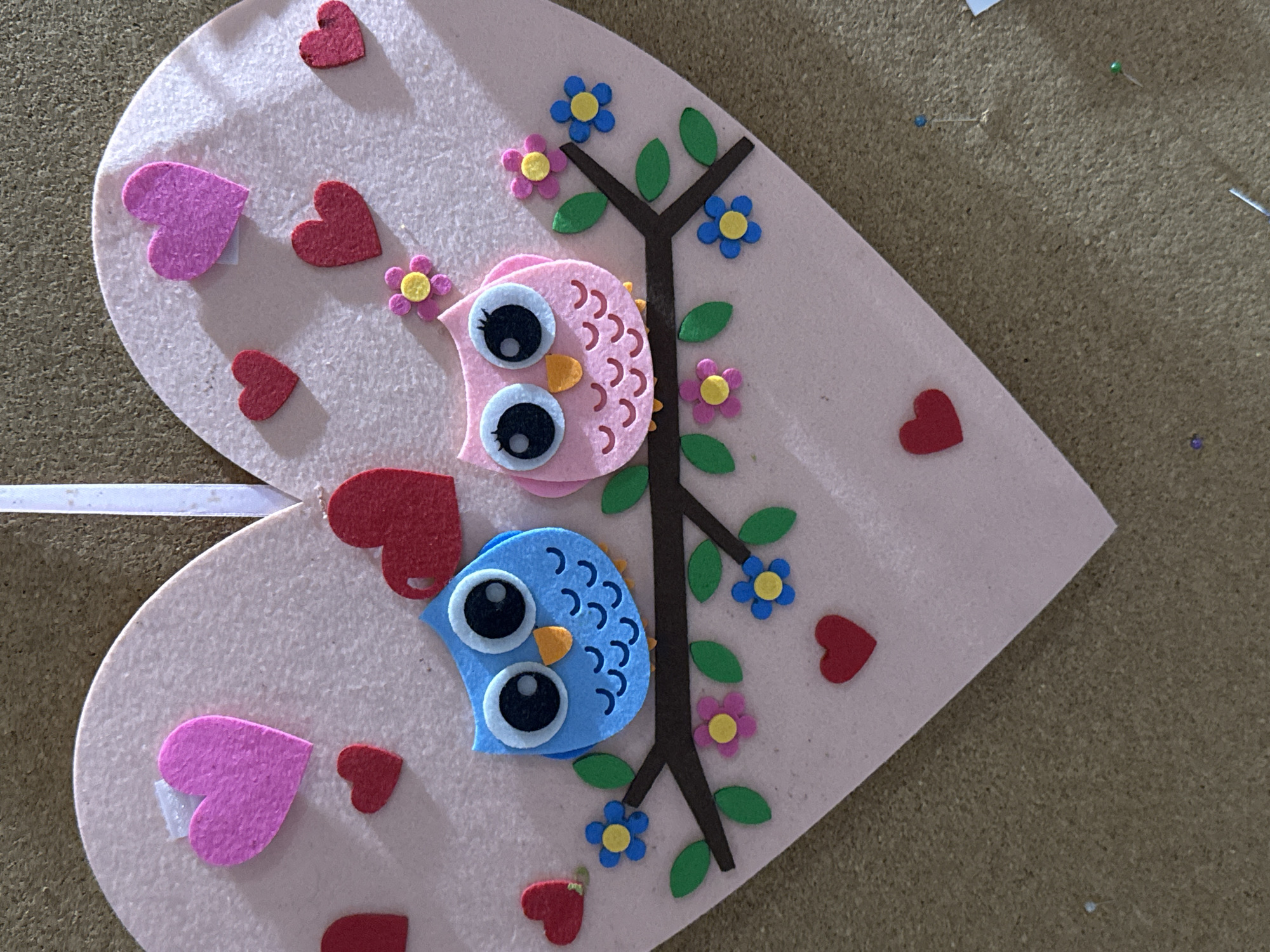 This media file features a close-up, high-angle shot of a heart-shaped craft made of light pink felt. The craft is adorned with two felt owls, one pink and one blue, positioned one above the other on a brown felt tree branch. The owls have large, black and white felt eyes, orange felt beaks, and detailed red and blue wavy lines on their chests, respectively. The tree branch is decorated with green felt leaves and small felt flowers in pink and blue with yellow centers. Scattered around the owls and tree are several small red felt hearts and two larger pink felt hearts. A white ribbon is attached to the left edge of the heart-shaped craft. The background appears to be a corkboard surface, with several small pins visible. The lighting suggests an indoor setting. No text is visible in the image.
