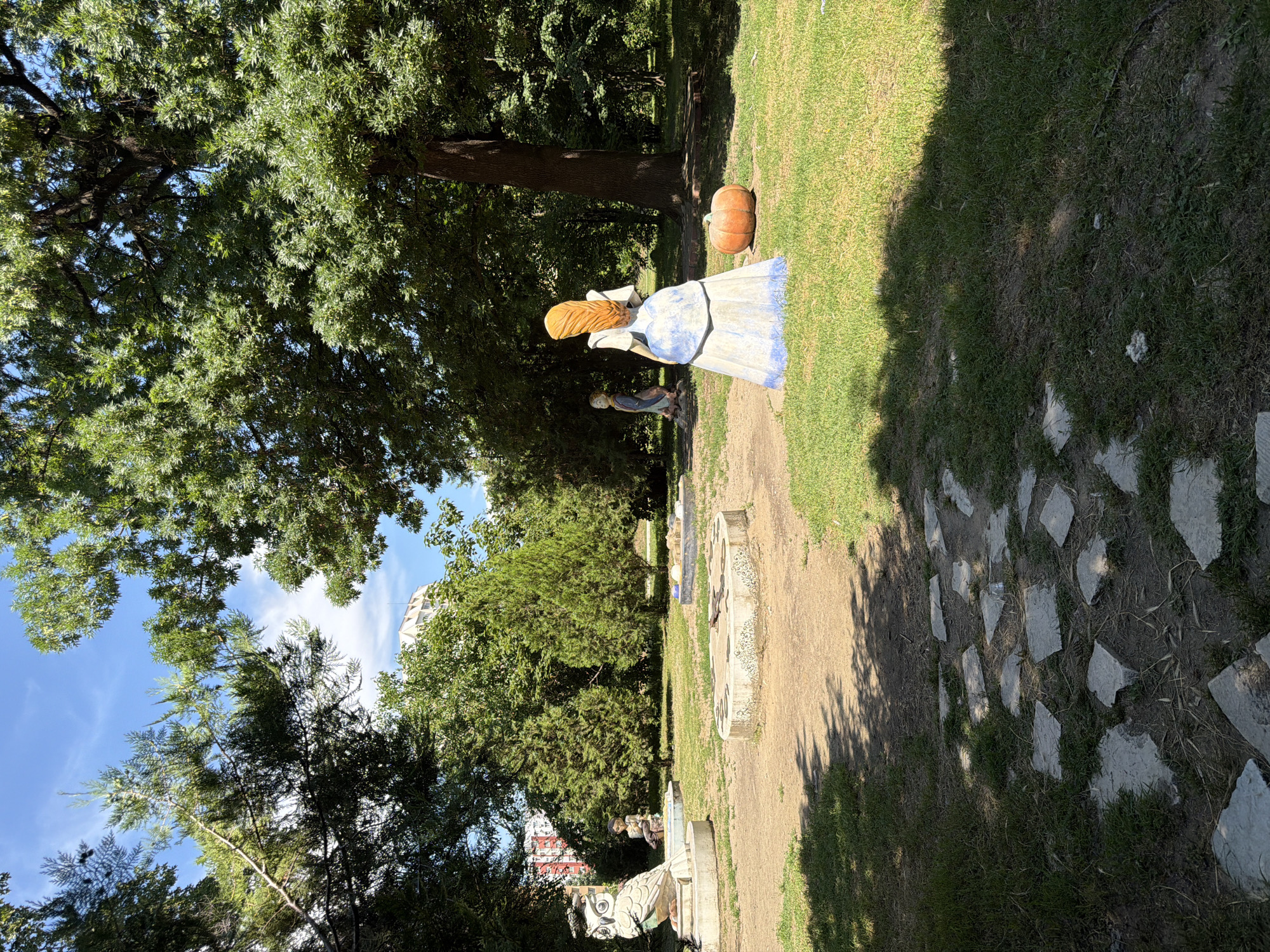 The image depicts a park scene in Pazardzhik, Bulgaria, featuring several concrete sculptures and natural elements. In the foreground, a pathway made of irregular flagstones winds through a grassy area dappled with tree shadows. To the right, a large, orange pumpkin rests on the grass next to a statue of a woman in a blue and white dress, her back to the viewer, with blonde hair. In the midground, a statue of an owl is visible on the left, with a fountain or decorative structure beside it. Further back, a small statue of a boy sits near a tiered, circular structure. The background is filled with lush green trees and foliage, with a building visible in the distance through the trees. The sky is blue with scattered white clouds. The overall impression is of a whimsical and somewhat overgrown public garden.