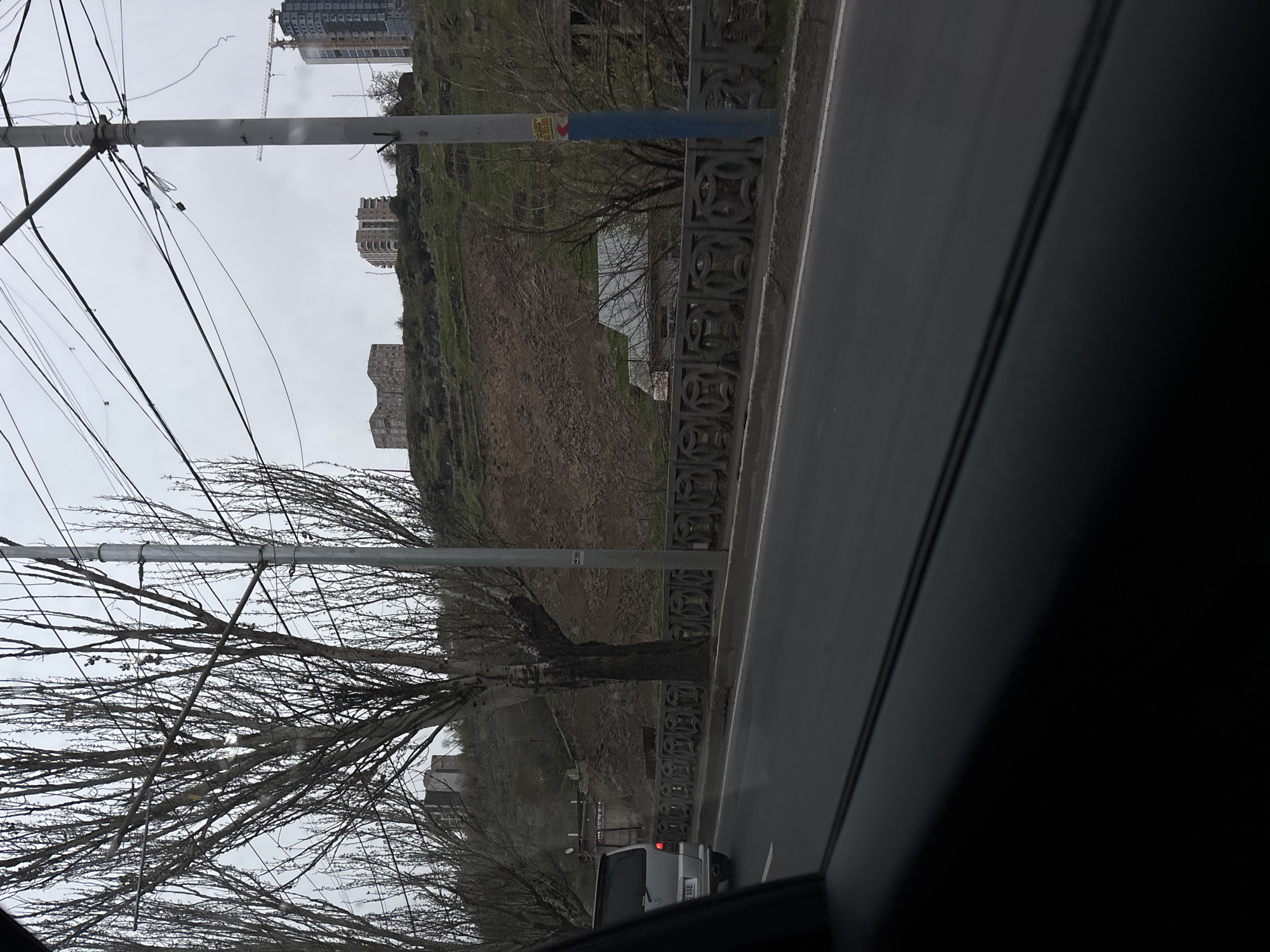 A view from a car interior shows a street in Yerevan, Armenia, with a hillside rising on the left side. The hillside is covered in brown dirt and some green vegetation, with a low concrete wall at its base along the roadside. Several buildings are visible on the hillside and further up the incline. Power lines and poles obstruct the view towards the upper left. Leafless trees are scattered throughout the scene. A gray car is visible to the right in the foreground. The sky is overcast.