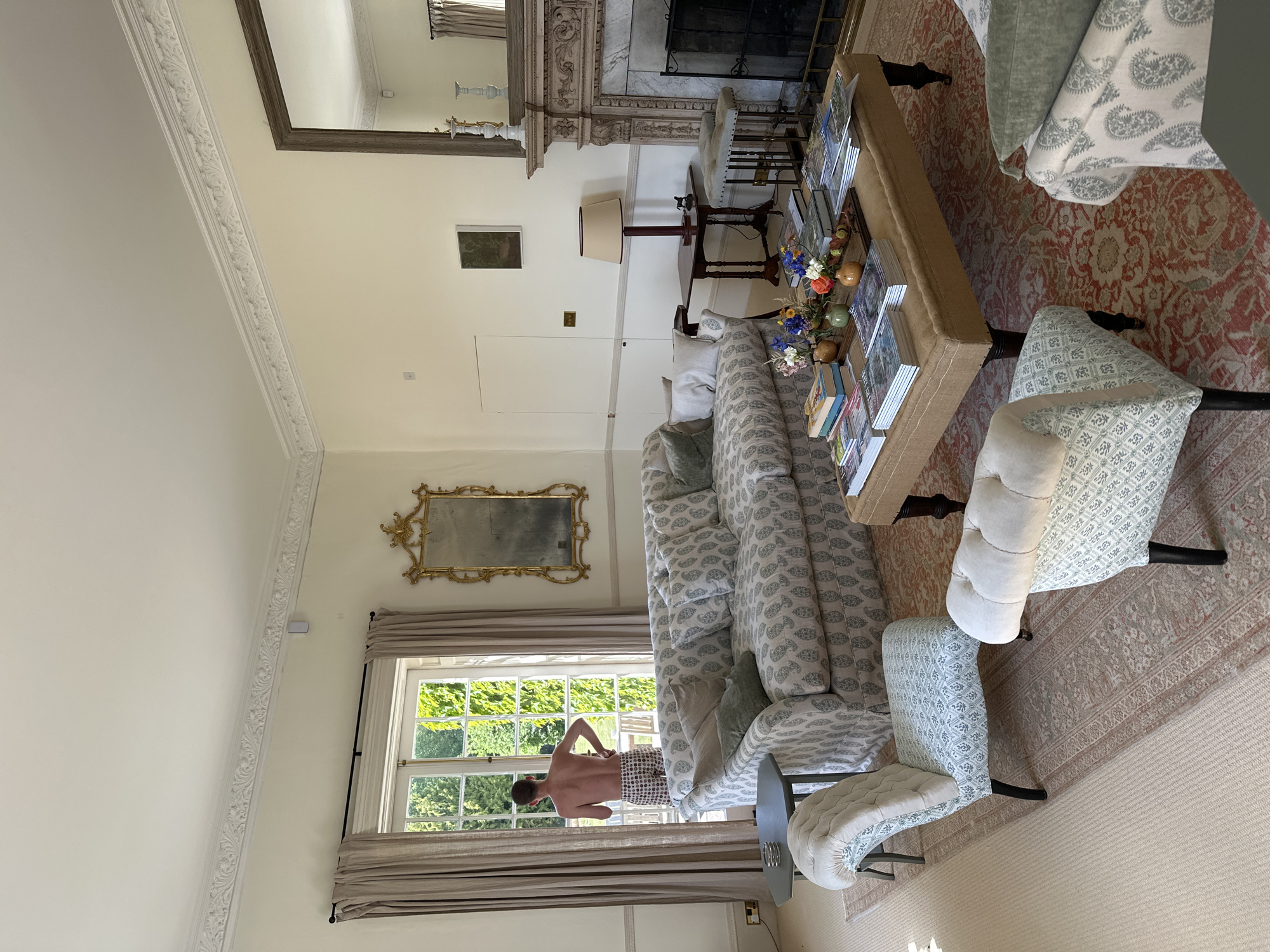 The image shows an interior view of a well-lit living room. The room has cream-colored walls and detailed crown molding. A large window is visible in the lower-left of the frame, partially covered by light beige curtains. A shirtless man is seen looking out the window. There is a sofa with a paisley pattern and several chairs with a similar patterned fabric and button-tufted backs. A long, rectangular coffee table laden with books and floral arrangements is in the center of the room. A large rug with a red and beige pattern covers part of the floor. A large, ornate mirror hangs above a fireplace with intricate stone detailing. A smaller framed picture hangs on the wall near a table lamp.