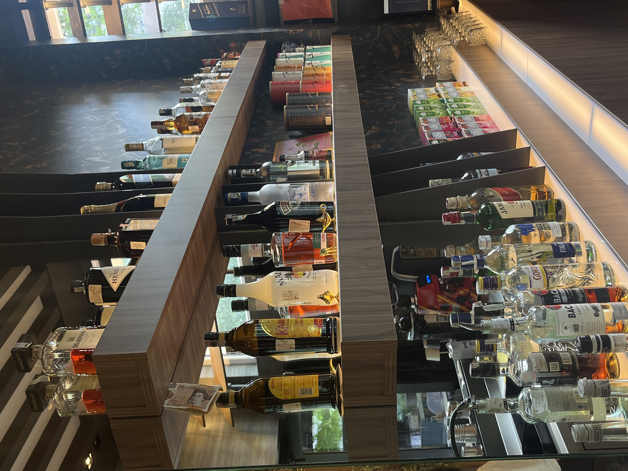 This image captures a wide-angle, eye-level view of a well-stocked bar counter and shelves within an establishment in Plovdiv, Bulgaria. The scene is devoid of people, activity, or interaction.
The focal point is a multi-tiered shelving unit crafted from wood-laminate, extending horizontally across the frame and displaying a diverse collection of alcoholic beverages.
**Upper Shelf:**
The topmost shelf features larger bottles, including a prominently displayed Disaronno liqueur bottle, another labeled "HENNESSY" (cognac), and a "MOËT & CHANDON" (champagne) bottle. To the far left, a bottle with red liquid is partially visible, and a folded banknote is tucked under the shelf's edge, partially obscuring its denomination. Various other spirits and liqueurs in glass bottles are arranged neatly.
**Middle Shelf:**
Below, the middle shelf showcases a variety of popular spirits. Recognizable brands include Absolut vodka, Grey Goose vodka, Bacardi rum, Malibu coconut liqueur, Kahlua coffee liqueur, and Aperol spritz. Some bottles feature small decorative items, such as a golden genie lamp figurine next to the Aperol bottle. This shelf is subtly illuminated from below by a concealed light strip, casting a warm glow on the bottles.
**Lower Shelf/Counter:**
The lowest level serves as the primary bar counter area. It holds numerous clear glass bottles, some likely pouring bottles with speed pourers, along with shakers and other bar tools. An espresso machine, identifiable by its red housing and blue display light, is positioned towards the center-right. Further right, stacks of inverted clear wine and cocktail glasses are meticulously arranged. Behind the glassware, multiple cartons of various colorful juices or mixers are neatly stacked.
**Background and Environment:**
The back wall behind the shelves is covered in dark wallpaper featuring a subtle, ornate pattern that appears to depict foliage or abstract designs. To the far right, a tall window provides natural light and a blurred view of green foliage outside, suggesting an outdoor garden or street scene. The overall lighting is a combination of artificial light from the bar area and natural light from the window, creating a well-lit but not overly bright ambiance. The floor and immediate foreground are not clearly visible. The establishment appears to be tidy and organized, indicating an operational commercial space.