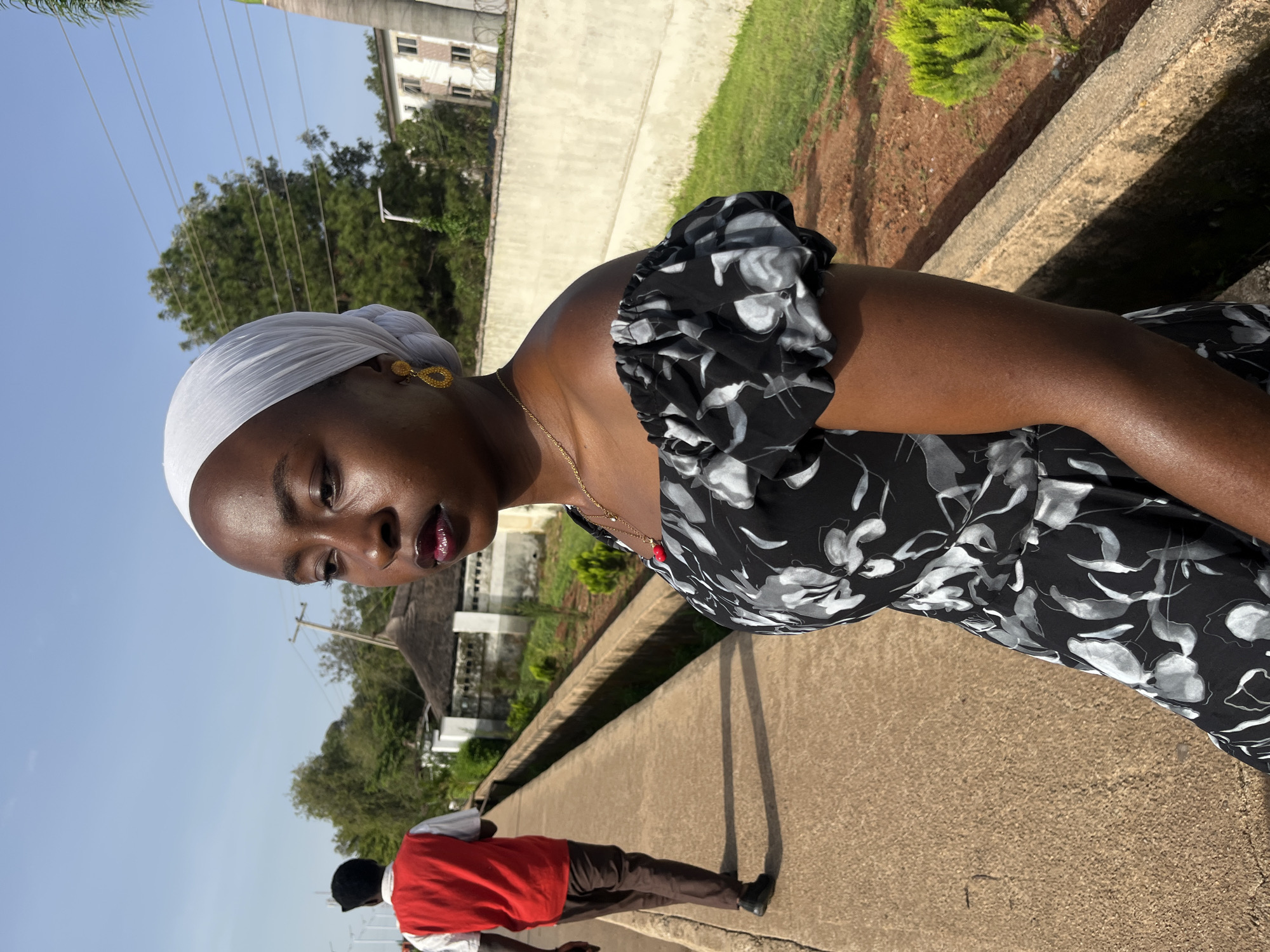 A medium shot captures a young Black woman from the chest up, facing to the left and looking away. She has dark skin, a white headscarf, and is wearing a black and white floral off-the-shoulder dress. Gold earrings and a delicate gold necklace with a red pendant adorn her. Her lips are painted with dark lipstick. In the background, a man in a red vest over a white shirt and brown pants walks away from the camera on a concrete path. The path is bordered by a raised concrete edge and a dry, dusty ground area with sparse green bushes and a thatched-roof structure. Beyond this, a concrete wall with a chain-link fence separates the area from a building with multiple windows and a tree. The scene appears to be set in Bukuru, Nigeria, under a clear blue sky with visible power lines.