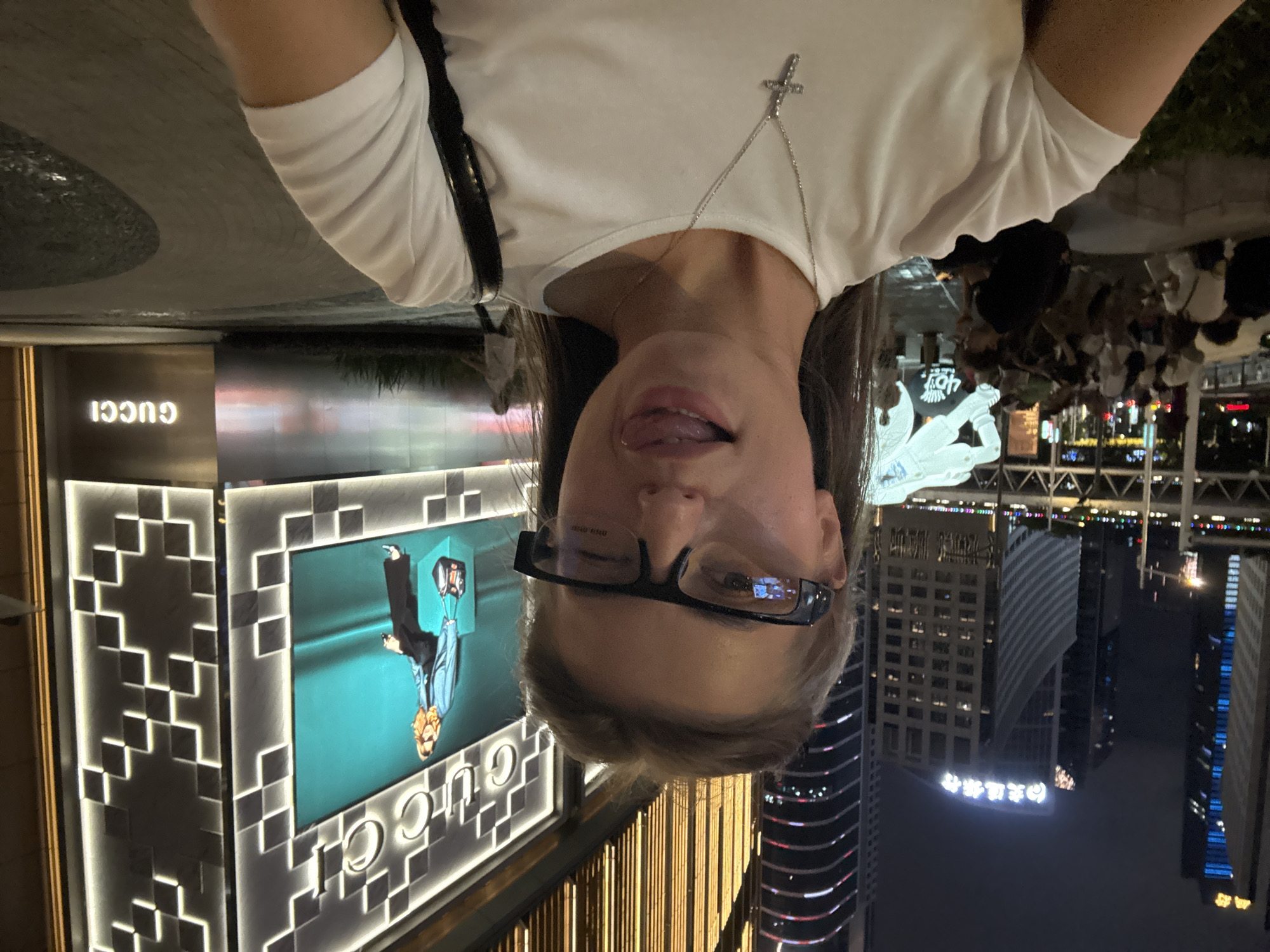 A selfie is taken at night in Tianhe District, China, in front of a Gucci store. The subject, a woman wearing glasses and a white t-shirt with a cross necklace, sticks her tongue out playfully. Behind her, a large Gucci advertisement featuring a blonde woman is displayed on a screen. The background shows illuminated buildings and a crowd of people gathered in an outdoor area, suggesting a public space or event. The environment is characterized by urban night lighting and modern architecture.