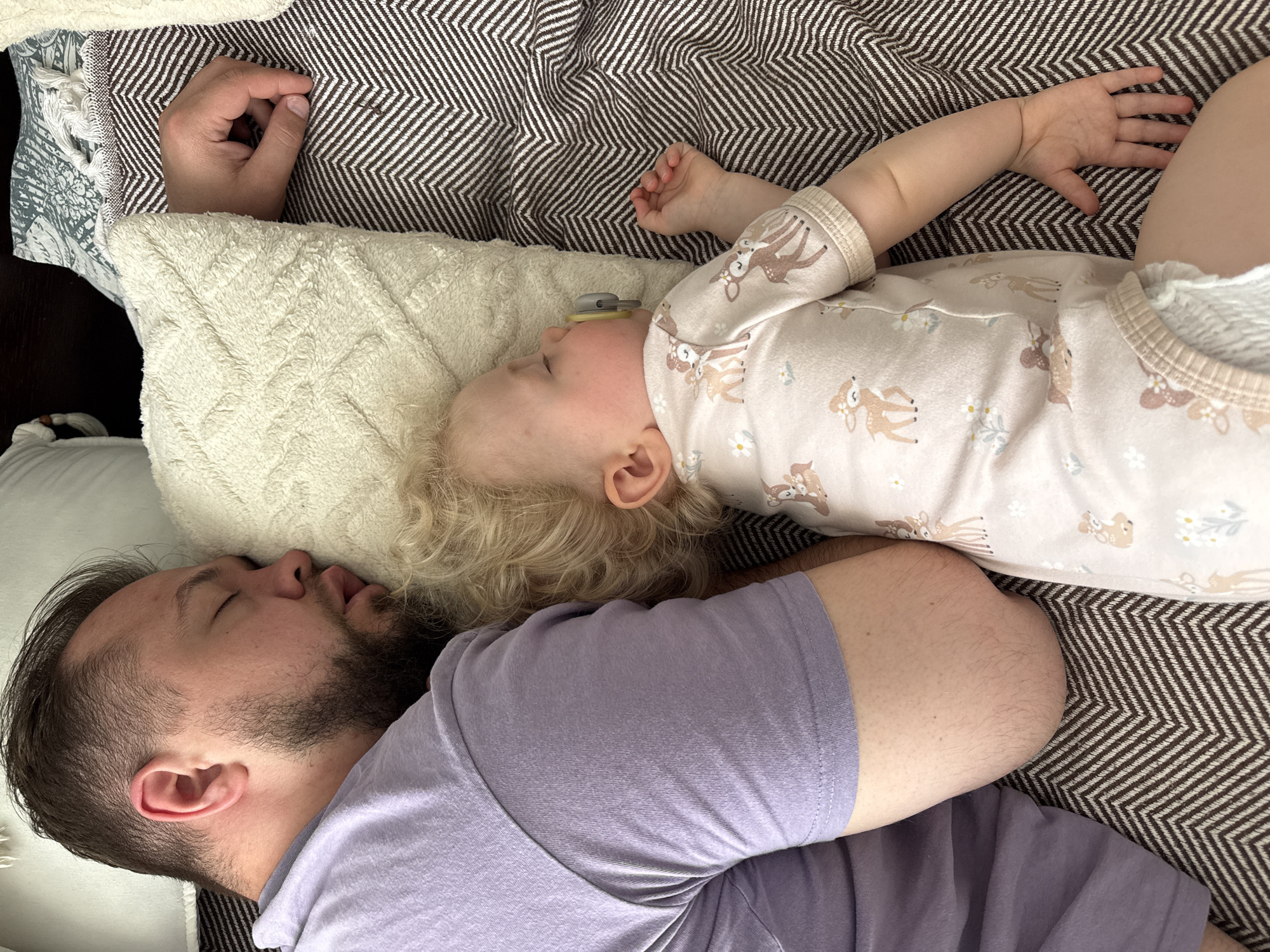 A man and a baby are sleeping on a bed. The man is lying on his side, wearing a purple t-shirt. The baby is lying on its back with a pacifier in its mouth and wearing a fawn-patterned onesie. The bedding consists of a white pillow and a brown and white chevron-patterned blanket. The location appears to be indoors. There is no indication of the location being in the city of Vladimir, Russia.