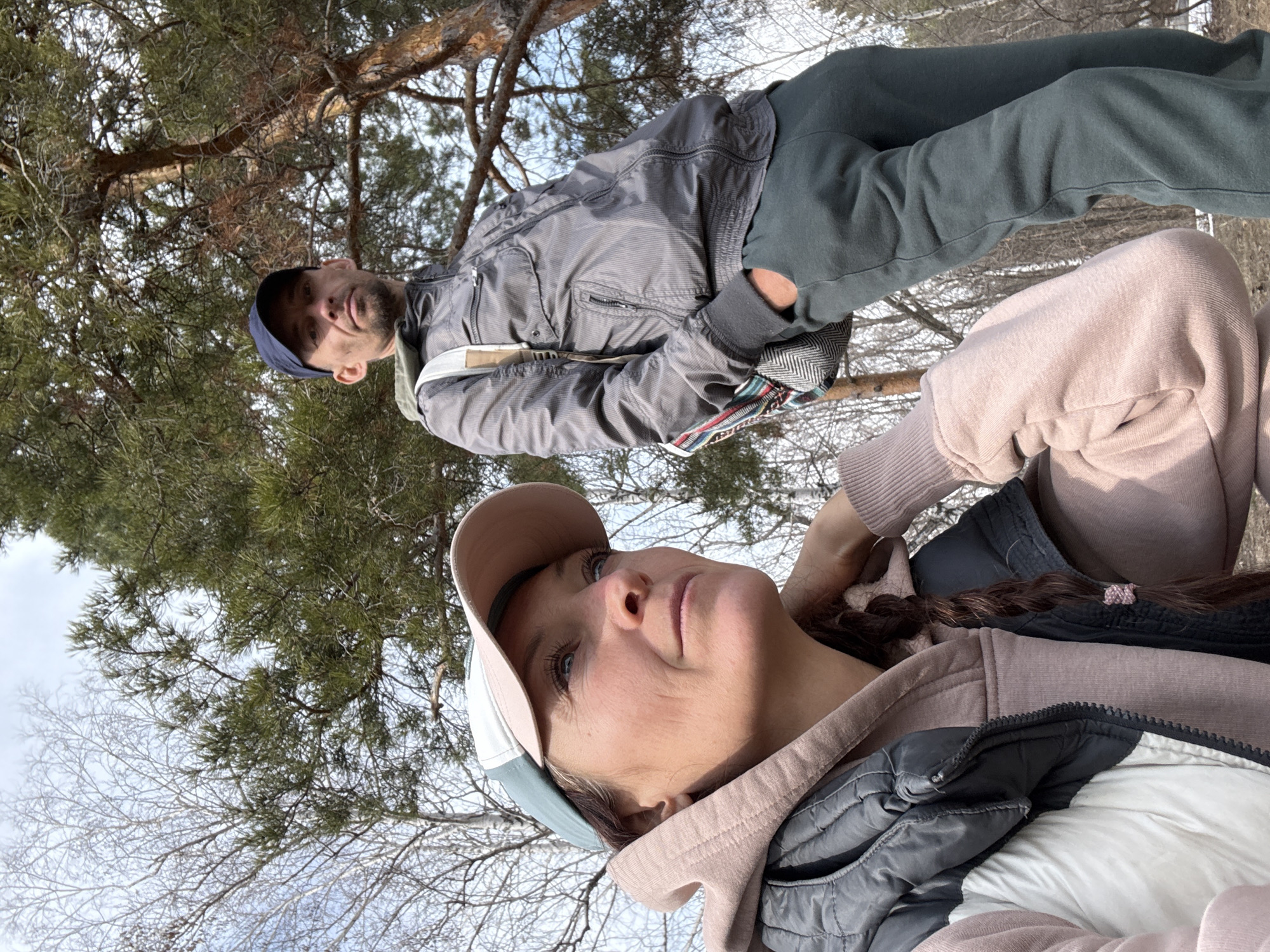 Two people are present in the image, seemingly taken outdoors. The background suggests a wooded area with trees and sky visible.
One person, a woman, is closer to the camera, wearing a baseball cap and a layered outfit consisting of a hoodie and a vest. She has braided hair and is looking upwards.
The second person, a man, is positioned further away in the background. He is wearing a jacket, possibly a light colored undershirt, dark pants and a head cover. He is looking towards the camera.
The setting is unclear from the image details, but the request specifies Ulyanovsk, Russia as the location. The wooded area suggests a park or forest location within or near the city.