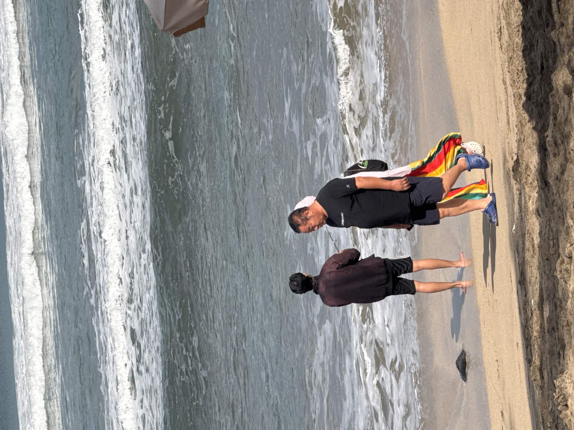 Two men are on a sandy beach in Kuta, Indonesia, facing the ocean. The man on the left wears a dark jacket and shorts, with his back to the camera, and is standing in the shallow water. The man on the right wears a black t-shirt and navy shorts, with a colorful cloth draped over his shoulder and arm. He is walking towards the right side of the frame and appears to be holding a fishing rod. Gentle waves are breaking on the shore. The sand is a light tan color, and there is a shadow of a beach umbrella visible in the upper right corner.