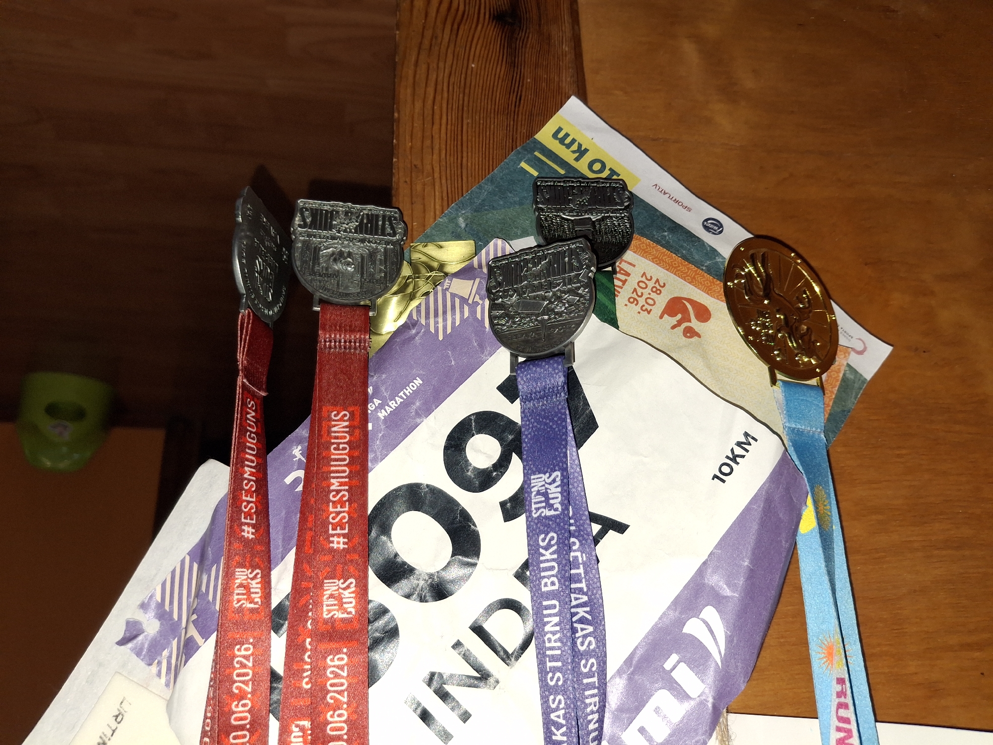 A close-up, eye-level shot shows multiple running medals hanging from a wooden surface, likely a wall or shelf. The medals are attached to brightly colored lanyards, including blue, purple, and red. Some lanyards have text and logos, such as "RUN", "STIRNU BUKSS", and "#ESESMUUGUNS". One prominent gold medal features an embossed image of a deer or elk. Several silver medals are also visible, each with intricate engravings depicting what appear to be scenes of nature or events. In the background, a large, crumpled piece of paper or banner with text like "10KM" and "MARATHON" is partially visible. A small, green, owl-shaped object, possibly a child's potty training seat, is visible in the upper right corner. The image is well-lit, with the wooden surface exhibiting a distinct grain pattern. The scene appears to be indoors, given the absence of natural light and the presence of the green object. The location of Ogre, Latvia, is not explicitly indicated in the image itself, but the text on the medals and banner might suggest a connection to events held there.