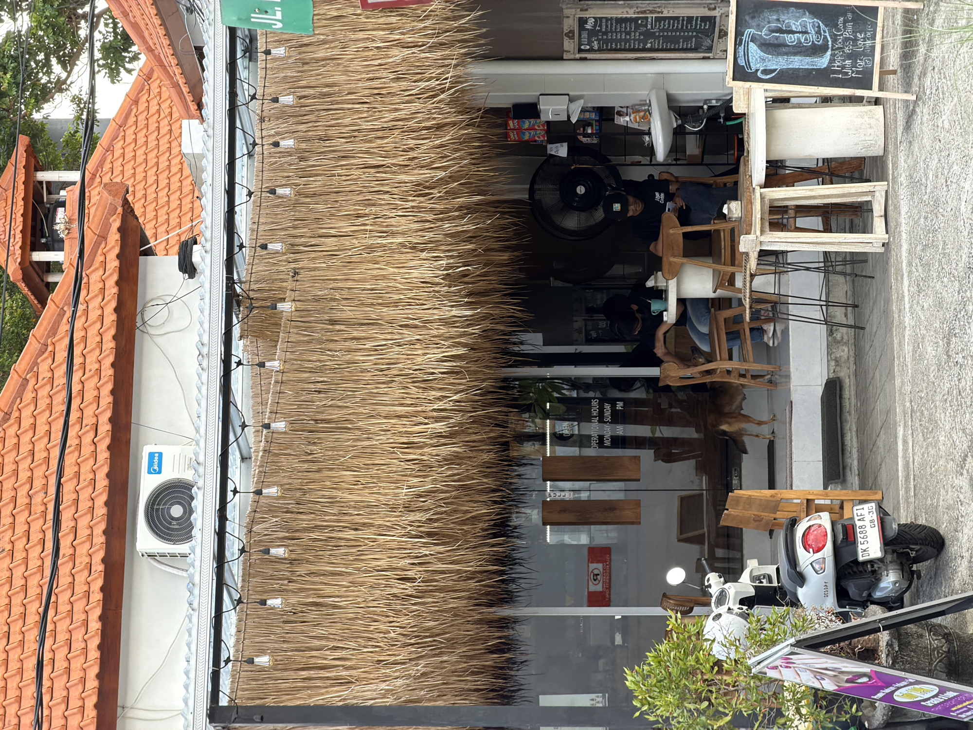 The image depicts the exterior of a cafe in Kuta, Indonesia, with a prominent thatched roof overhang. In the foreground, a white scooter with license plate DK 5688 AF1 is parked on the pavement. A purple A-frame sign is leaning next to the scooter. Behind the scooter, an open-air cafe seating area is visible through a glass storefront. Inside, two individuals are seated at tables, one with a blue mug and the other looking at their phone. A large fan is positioned behind them. A dog is standing in the doorway. A chalkboard sign with a drawing of a hand holding a coffee cup and text that reads "I hope you come with less pain at More Coffee" is placed on the pavement. The cafe has an air conditioning unit mounted on the wall above the thatched overhang, and decorative string lights are suspended from a metal frame. The tiled roofs of adjacent buildings are visible in the background.