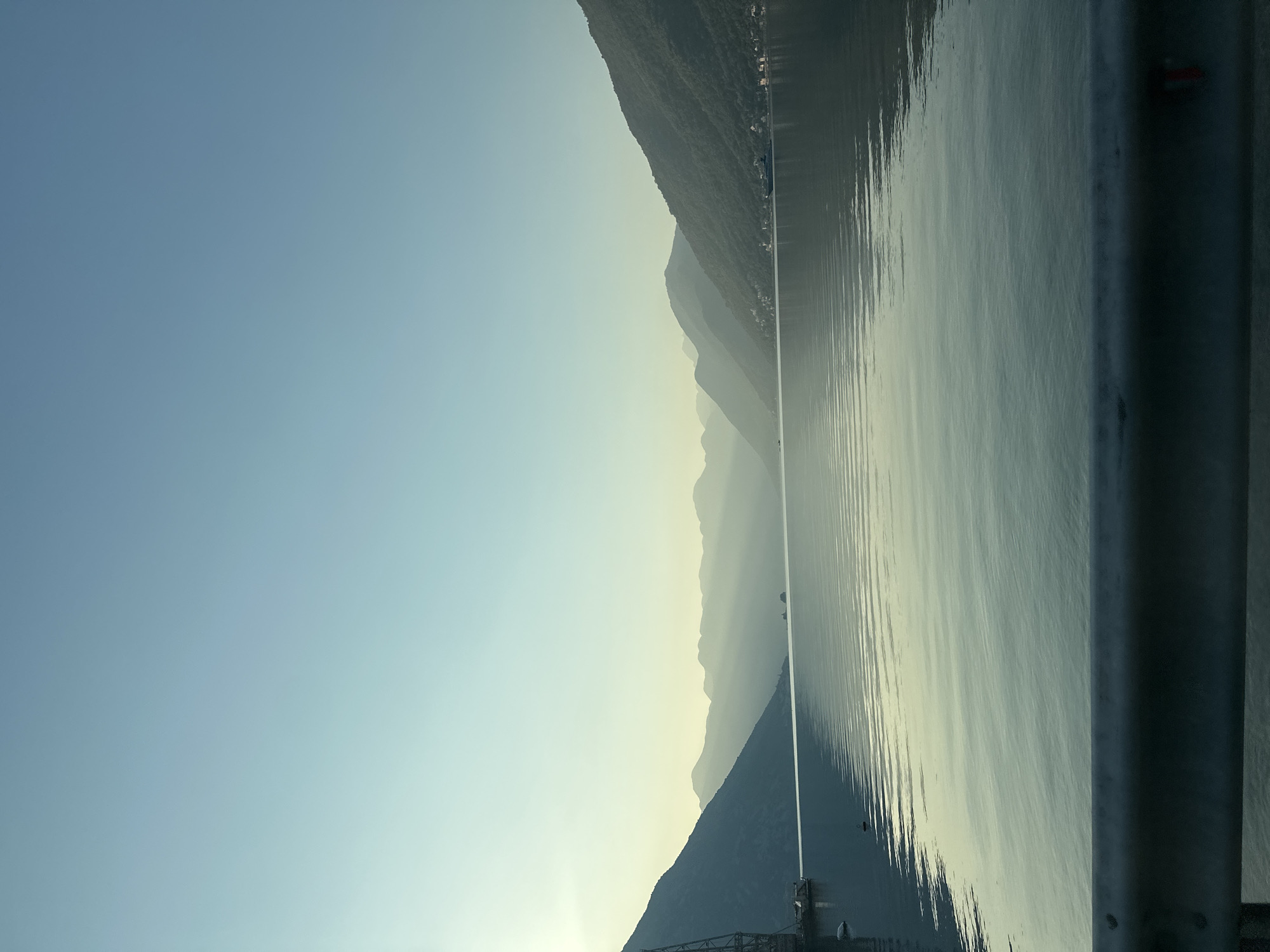 A wide-angle, vertical, eye-level shot shows a serene bay at Lipci, Montenegro, bathed in the soft, hazy light of early morning or late afternoon. The calm, rippling water occupies the lower half of the frame, reflecting the pale blue sky and the silhouettes of distant, layered mountains. The mountains, covered in lush green vegetation, form a dramatic backdrop, with sunlight piercing through the gaps between them, creating ethereal beams that stretch across the water.
On the left side of the bay, a tall, industrial-looking structure, possibly a crane or a tower, is partially visible, with a small boat moored nearby. On the right side of the bay, a narrow strip of land with scattered houses and buildings is nestled at the foot of the mountains, suggesting a small settlement. A large ship or ferry is visible in the distance on the right, moving across the water. The foreground is dominated by a metal guardrail, its textured surface adding a layer of urban or infrastructural element to the otherwise natural landscape. The overall atmosphere is peaceful and tranquil, emphasizing the grandeur of nature and the quiet coexistence of human activity within it.