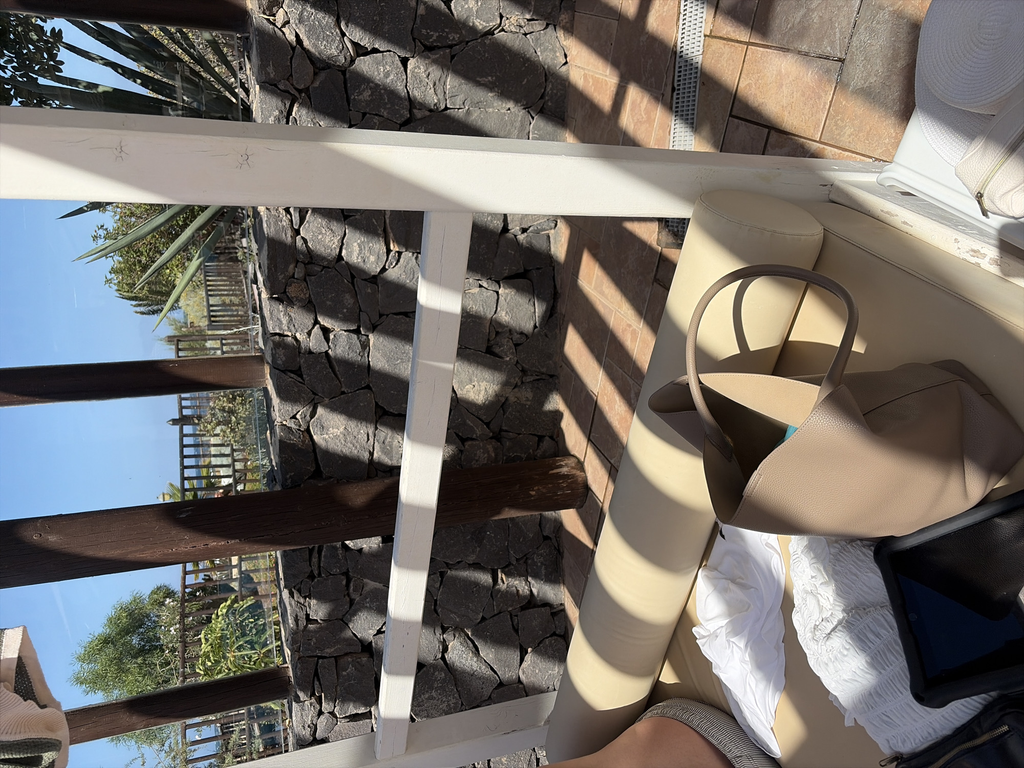 This is a medium-angle, eye-level, outdoor shot taken in Antigua, Spain, on a sunny day. The scene depicts a lounge area with a light-colored cushioned daybed or seating area. Several items are placed on the daybed, including a beige tote bag, a white crumpled garment, and a tablet in a black case. To the left of the frame, a portion of a person's leg is visible, wearing a striped shoe or sandal.
The background features a dark stone wall and wooden posts supporting a white structure. Beyond the wall, a wooden fence overlooks a landscape with trees, vegetation, and a glimpse of the ocean under a clear blue sky. Sunlight casts strong shadows on the stone wall and the tiled ground of the lounge area, indicating a bright, sunny environment. The overall atmosphere suggests a relaxed, holiday setting in a coastal location.