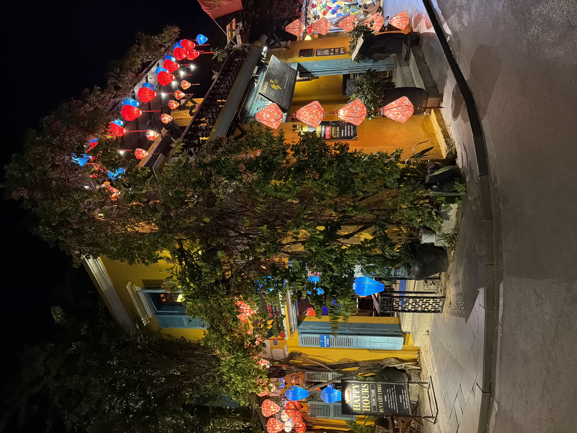 The image shows a street scene at night in Hội An Ward, Vietnam, featuring a yellow building illuminated by various colorful lanterns. The building has blue shutters on its windows and a balcony adorned with red and blue lanterns. A tree with dense foliage is positioned in front of the building, also decorated with lanterns. A sign on the left side of the image displays "HAPPY HOURS" with further details about a "BUY 1 GET 1 FREE" offer. Potted plants are visible near the entrance of the building, and the street in front is wet, reflecting the lights. The overall atmosphere is festive and vibrant, with the warm yellow of the building contrasting with the colorful lanterns and the dark night sky.