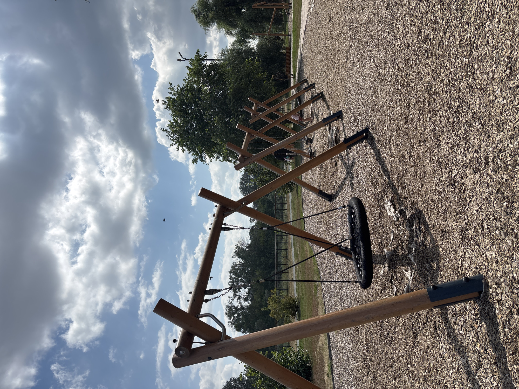 The image shows an outdoor playground with multiple swings made of natural wood structures. The ground is covered in wood chips. In the background, there are trees and a blue sky with scattered clouds. There are a few people visible in the distance, some of whom appear to be children. The scene is well-lit, suggesting it is daytime. There is no indication of the location being in Orenburg, Russia.