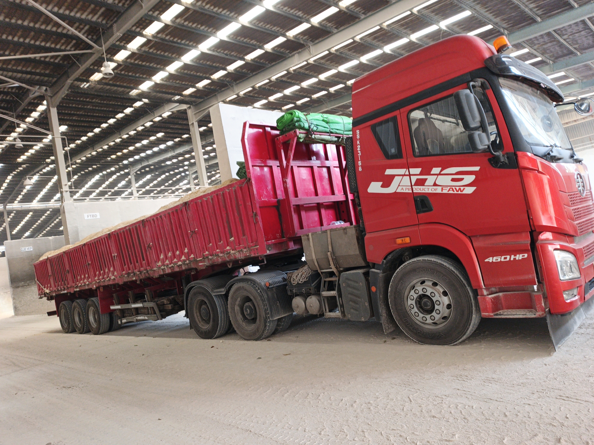 In Ngoro, Indonesia, a long, red FAW truck with a trailer filled with granular material is parked inside a covered industrial building. The building's roof is constructed of corrugated metal panels with numerous translucent skylights, allowing natural light to illuminate the interior. The truck has a large, red cab with "JHG A PRODUCT OF FAW 460HP" branding. The trailer is also red, with multiple axles and wheels, and is covered in green tarpaulins. The ground is a dusty, light-colored surface. The scene appears to be a loading or unloading area within a storage facility.