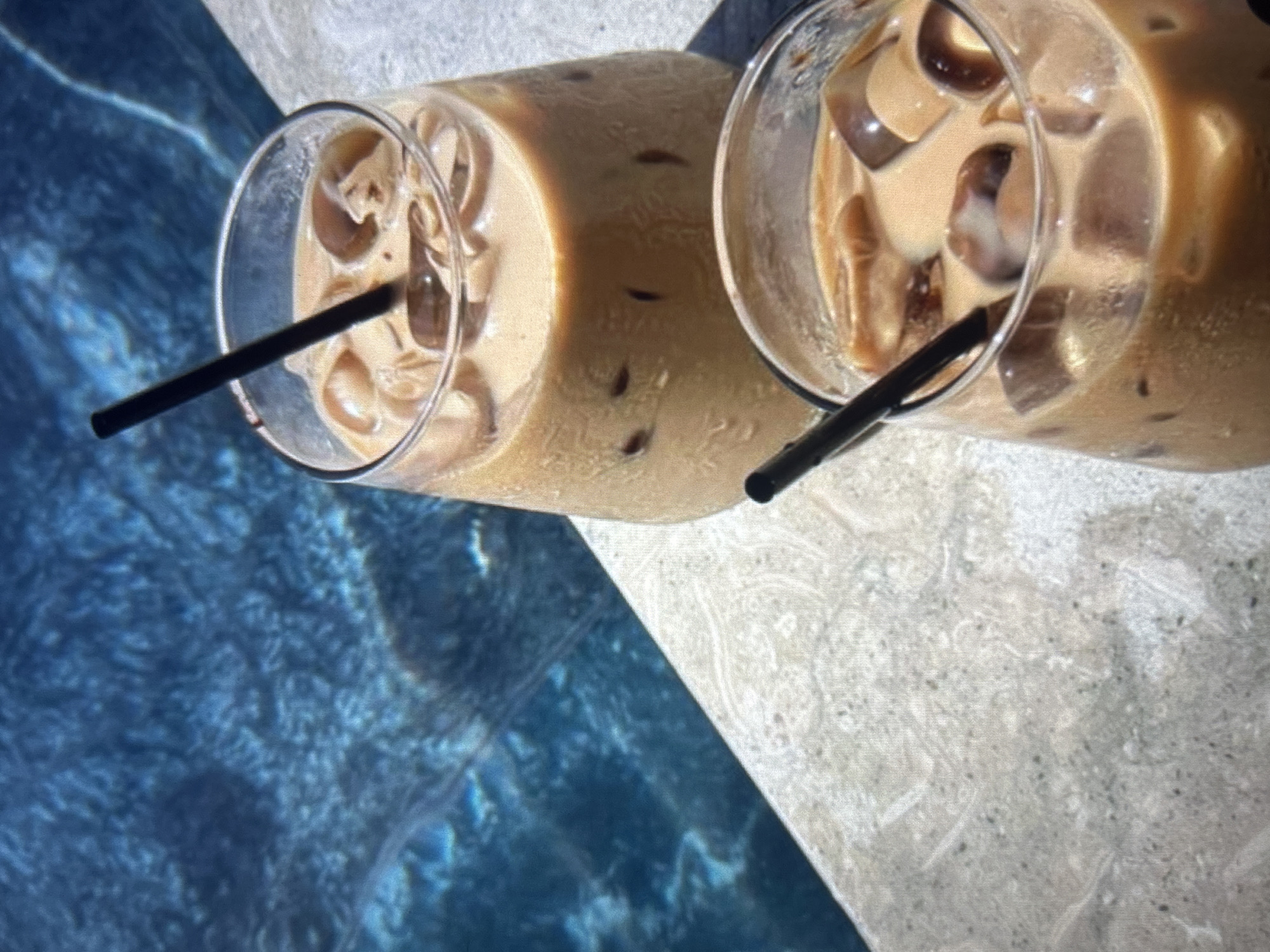 Two glasses of iced coffee sit on the edge of a swimming pool. The pool water is a deep blue, with sunlight reflecting off its surface. The glasses are clear and filled with a light brown liquid and ice cubes. Black straws protrude from each glass. The poolside edge is a light-colored, textured material. The scene is brightly lit, suggesting it is daytime. The specific location in Knezha, Bulgaria, is not determinable from the visual information provided.