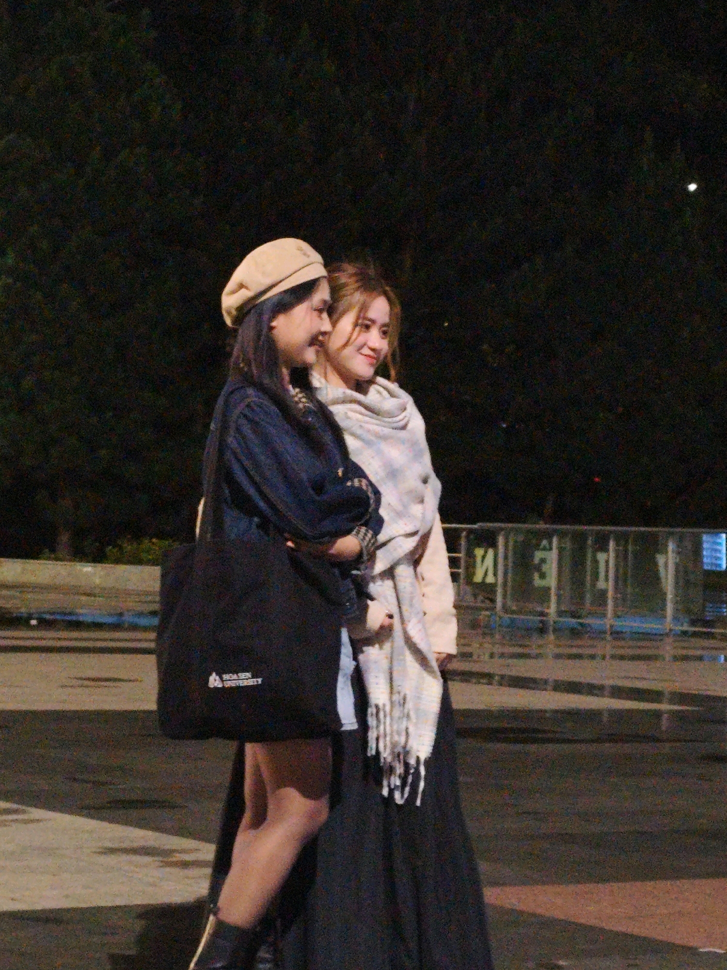 Two young women stand in profile, facing right, against a dark background of trees and a fence. The woman on the left wears a tan beret, a dark denim jacket, and a long black skirt. She carries a black tote bag with a logo that reads "HOA SEN UNIVERSITY." The woman on the right wears a light-colored scarf draped around her neck and a pale pink cardigan over a dark dress. Both women are smiling and appear to be interacting with each other. The ground is paved with a mix of asphalt and concrete, with some debris scattered on the surface. In the background, a metal fence with some lettering on it is visible, and behind that, there are some blurry lights. The scene appears to be outdoors, possibly at night, in an urban setting in Da Lat, Vietnam.