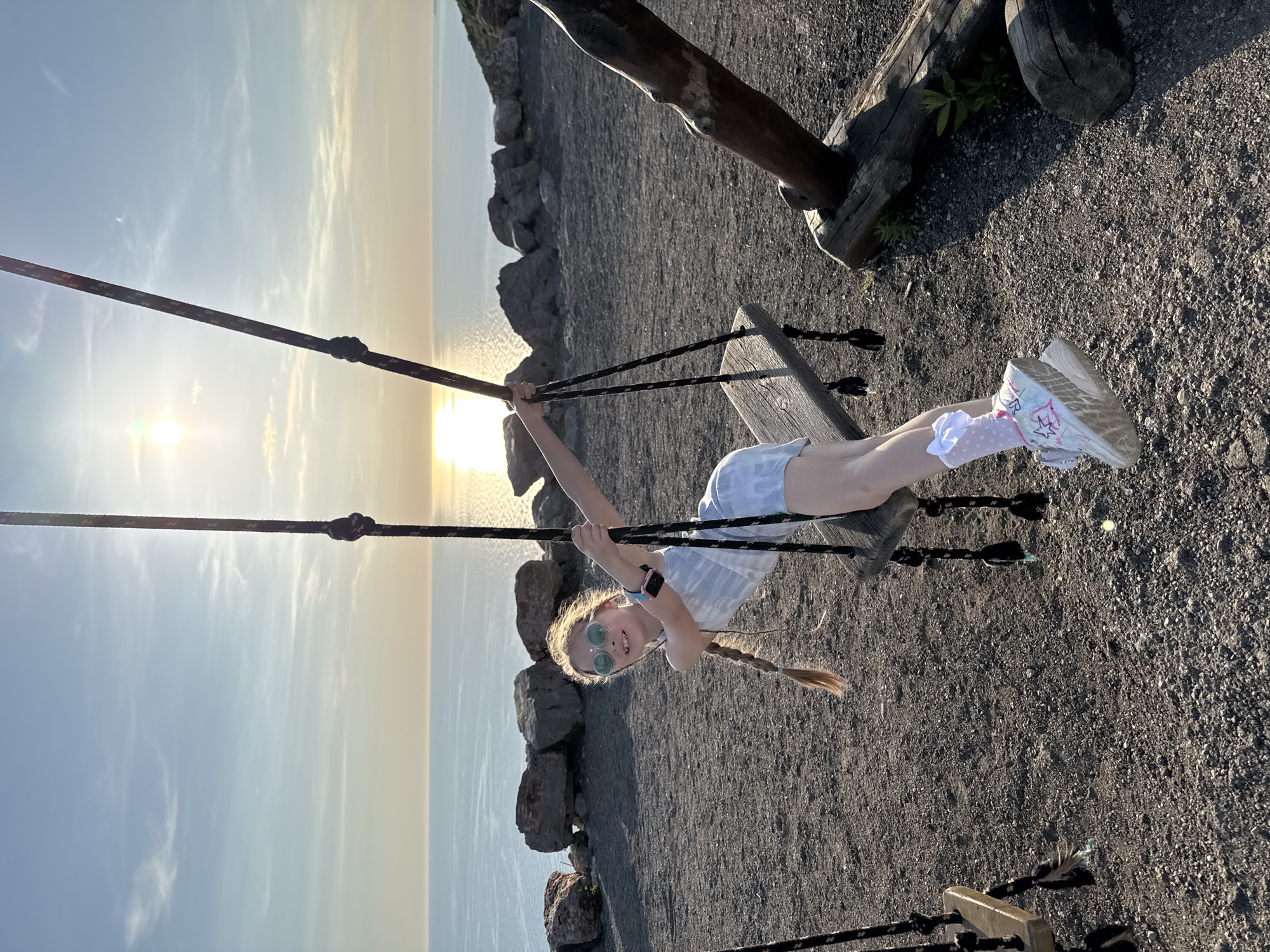 This outdoor photograph, taken at sunset, depicts a young girl joyfully swinging with a vast body of water in the background. The low sun, positioned on the left, casts a bright glare over the water and bathes the scene in warm, golden light.
The central figure is a young girl with blonde hair in a long braid. She wears light-colored sunglasses, a striped t-shirt, shorts, white polka-dotted socks, and white sneakers decorated with colorful, graffiti-style drawings including stars and squiggles. A pink smartwatch is visible on her left wrist. She is actively on a wooden swing, holding the ropes with both hands, leaning back with her legs extended, and smiling broadly at the camera.
The swing's ropes are dark and appear sturdy, attached to a rustic wooden frame, parts of which are visible on the right. The ground is covered in dark gravel or small stones. Beyond the swing and the girl, a rocky embankment or cliff edge leads down to a wide expanse of water, likely a sea or large lake, stretching to the horizon. The sky is clear with delicate, wispy clouds, glowing intensely near the sun. Another substantial wooden log structure lies on the gravel to the right.
The image captures a happy, playful moment against a beautiful, serene backdrop, likely a coastal or large lake environment in Kovačko Polje, Montenegro, as indicated by the context. No explicit text is visible.