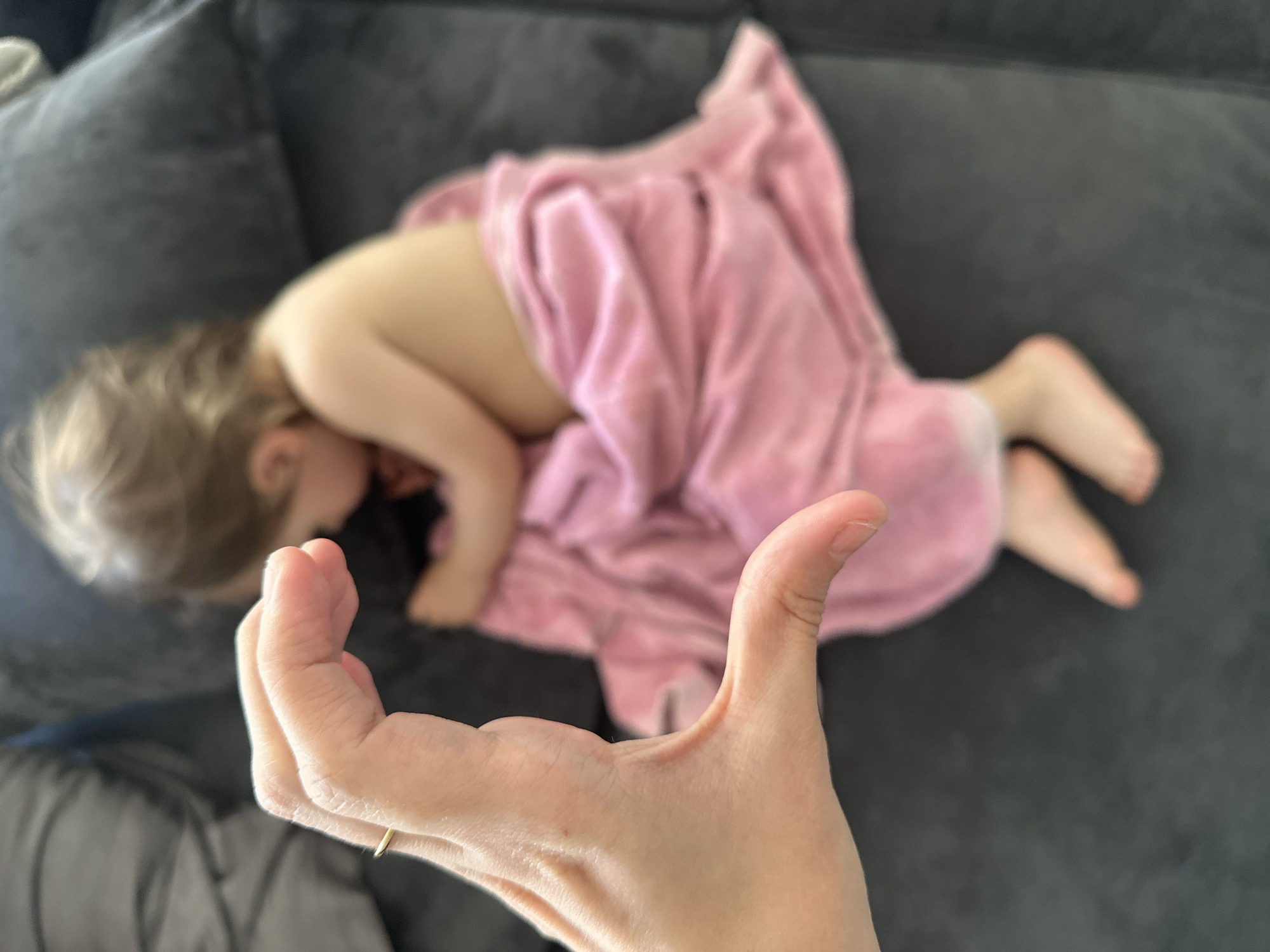 A child is sleeping on a gray couch covered by a pink blanket. A human hand is in the foreground. The location is not evident from the image.