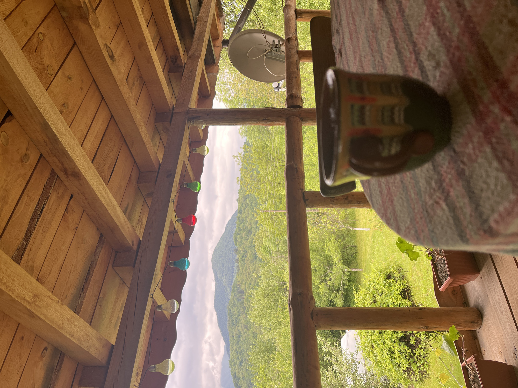 The image depicts an outdoor scene, seemingly from a porch or balcony, with a view of a forested landscape and a partial view of a mountain range. A wooden structure with visible rafters and a string of light bulbs, some colored, is positioned on the left. A wooden railing or ladder-like structure is in the center. A partially visible ceramic mug rests on a table covered with a patterned cloth. A satellite dish is visible outside, slightly above the wooden structure. The view outside shows a green landscape with trees. The location is assumed to be Shopite, Bulgaria. The image contains no visible people, interactions or actions.