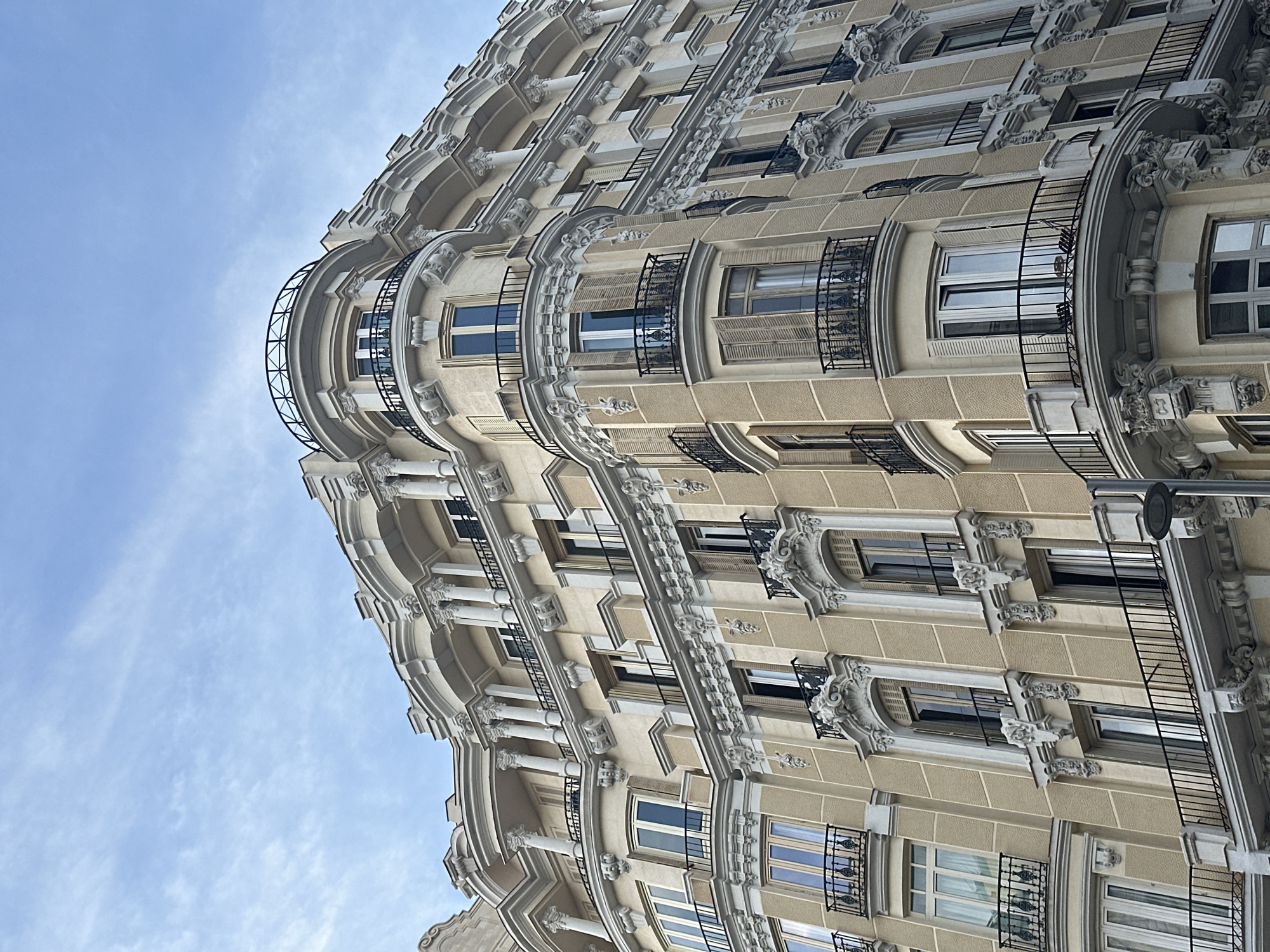 The image shows a low-angle view of a building in Madrid, Spain. The building has a rounded corner and is adorned with ornate architectural details, including balconies with black wrought-iron railings, decorative moldings, and classical-style columns. The building is light tan in color. The sky is visible in the background, with scattered cirrus clouds. A streetlight is visible in the lower-right corner.