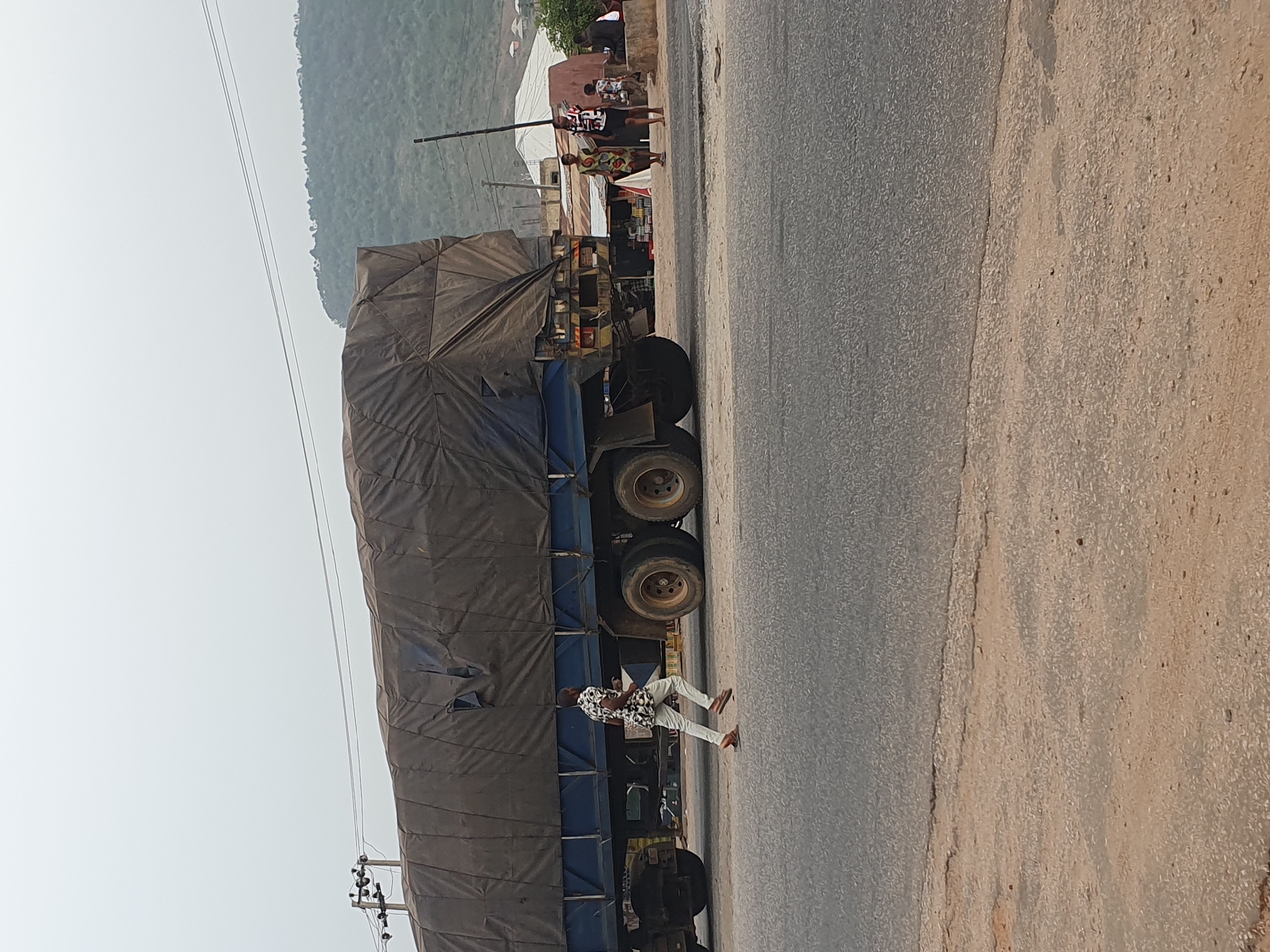 An outdoor scene captures a roadside in Lokoja, Nigeria, under bright daylight. The main subject is a very long, multi-axle truck, painted blue and heavily laden, with its cargo entirely covered by a dark, possibly black or dark blue, tarpaulin. The truck is positioned on the left side of the frame, seemingly parked or stopped by the side of a paved road.
To the immediate right of the truck, a man is walking from left to right, dressed in a patterned short-sleeve shirt, light-colored trousers, and sandals. Further along the road and behind the truck's cab, several other individuals are visible near small roadside stalls or shops, suggesting market activity. One woman in a patterned dress is clearly seen among them. The stalls display various goods, though specific items are not distinctly visible.
The paved road occupies the central and right portions of the image, appearing somewhat worn, with a distinct lighter-colored dirt or gravel shoulder to its far right. In the background, a large, undulating hill or mountain range stretches across the horizon, appearing hazy, indicating a clear but possibly dusty or humid atmosphere. Overhead, utility lines traverse the sky, connected to poles that are partially visible. The overall impression is one of a busy, functional roadside environment in a semi-urban or rural setting. No discernible text is visible on any objects or signs.