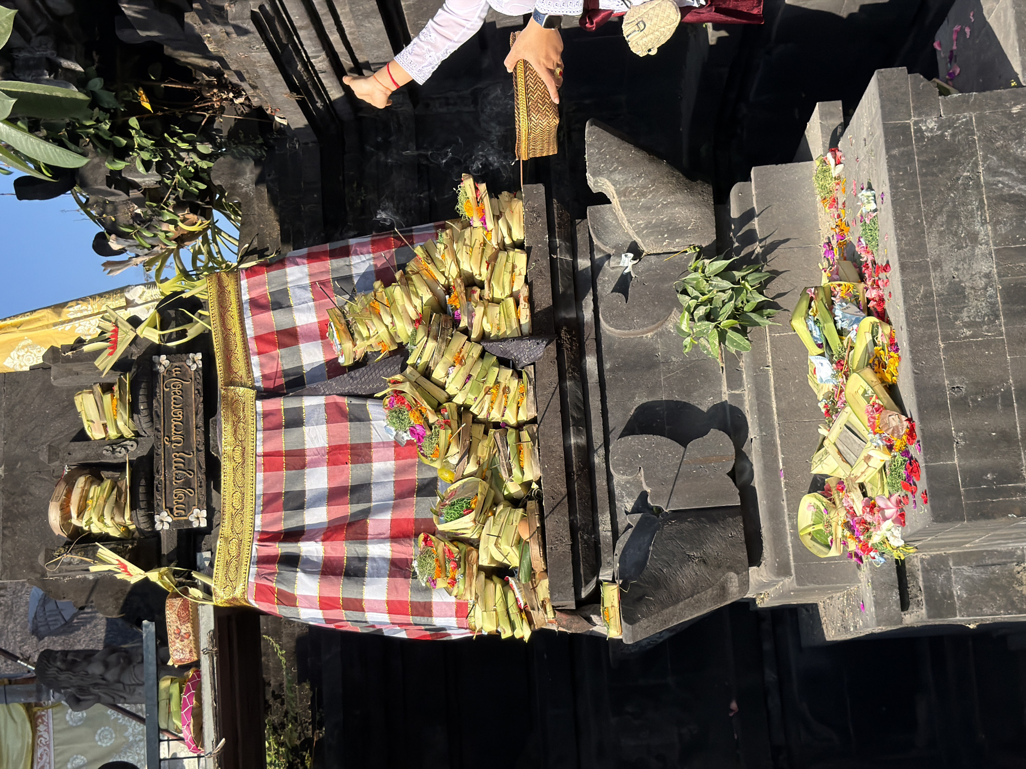 In the city of North Kuta, Indonesia, a vibrant outdoor scene unfolds at a stone altar adorned with offerings. A person, their arm outstretched and wearing a white lace top and a red sarong, holds a woven basket and gestures upwards as smoke rises from an incense stick. The altar is elaborately decorated with a checked cloth and stacked offerings of woven palm leaf containers filled with flowers, leaves, and what appears to be rice. More offerings, including scattered flower petals, lie on a lower level of the altar. The background features dark stone structures and lush green foliage, with a bright blue sky overhead.