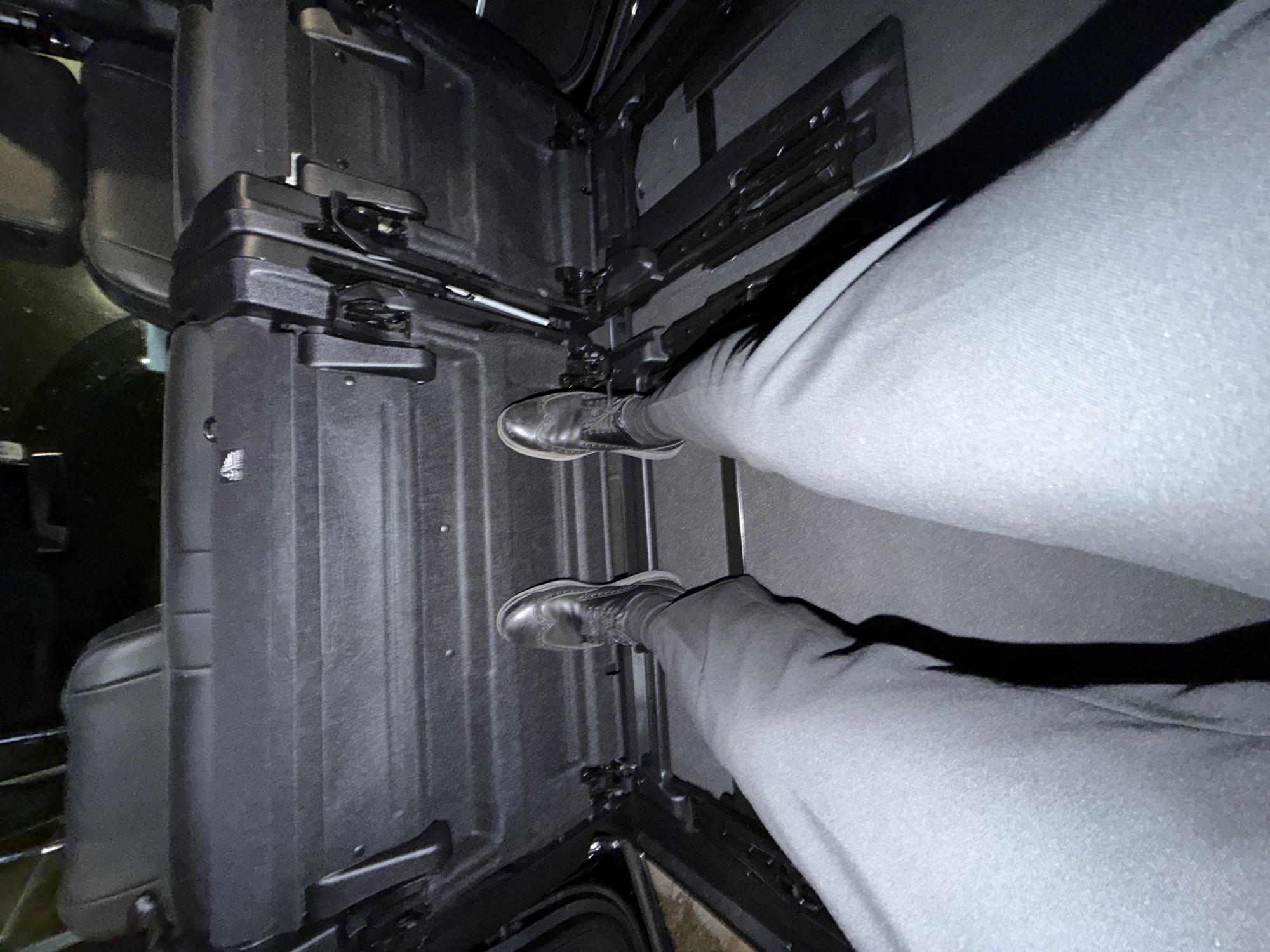 The image shows a close-up view of a person's legs and feet inside a vehicle, possibly a car or van, with the focus on the interior space. The person is wearing dark dress shoes and light-colored pants. The interior appears to be made of dark, textured plastic materials, with visible compartments, latches, and metal rails. The lighting is dim, suggesting it might be indoors or nighttime, and there are some shadows cast on the surfaces. The scene is viewed from a high angle, looking down at the person's legs and feet. The environment appears utilitarian and functional, likely part of the cargo or seating area of a vehicle. There is no visible text in the image. The setting is the Village of Southampton, United States of America.