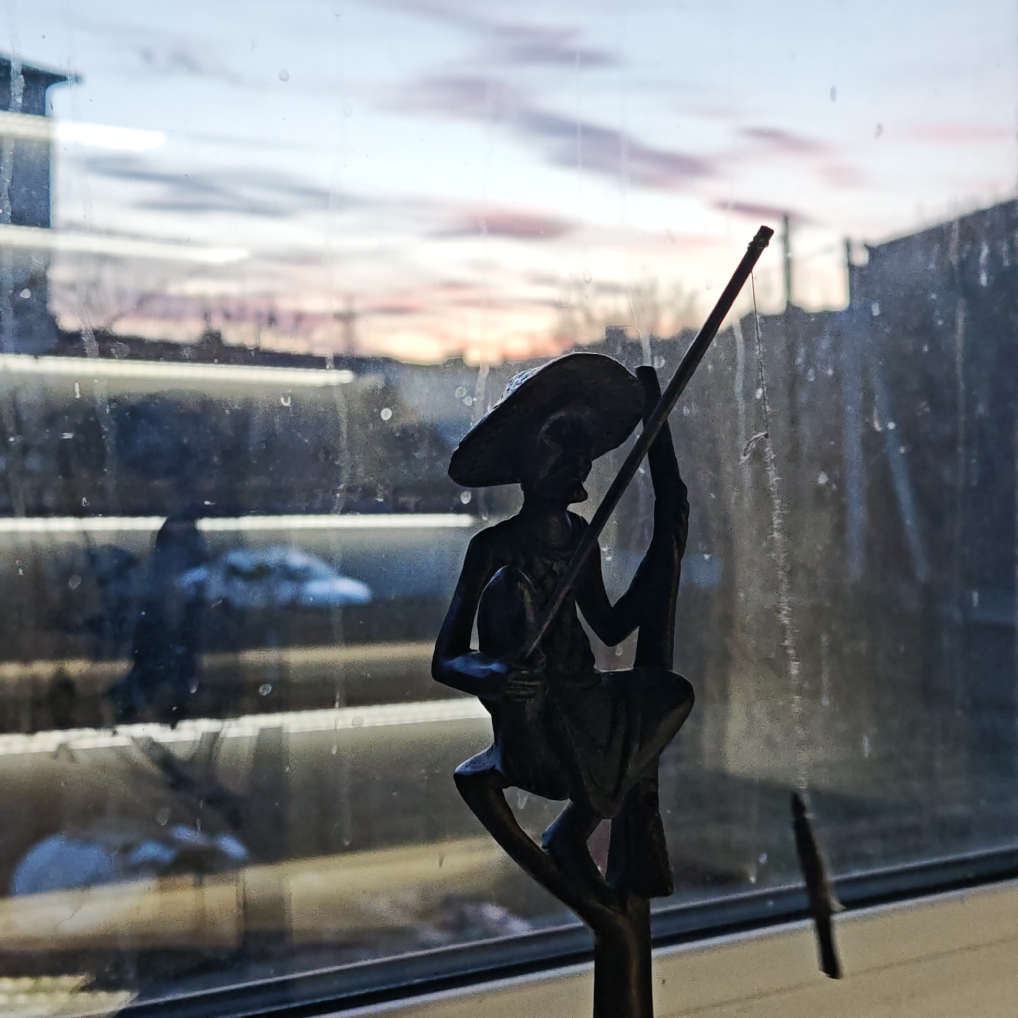 A figurine of a man with a hat, seated and holding a fishing pole, is placed on a windowsill. The window is streaked with grime. Outside, there is a building with horizontal blinds, and above the building, a cloudy sky with a hint of pink is visible. The location is Rostov, Russia.