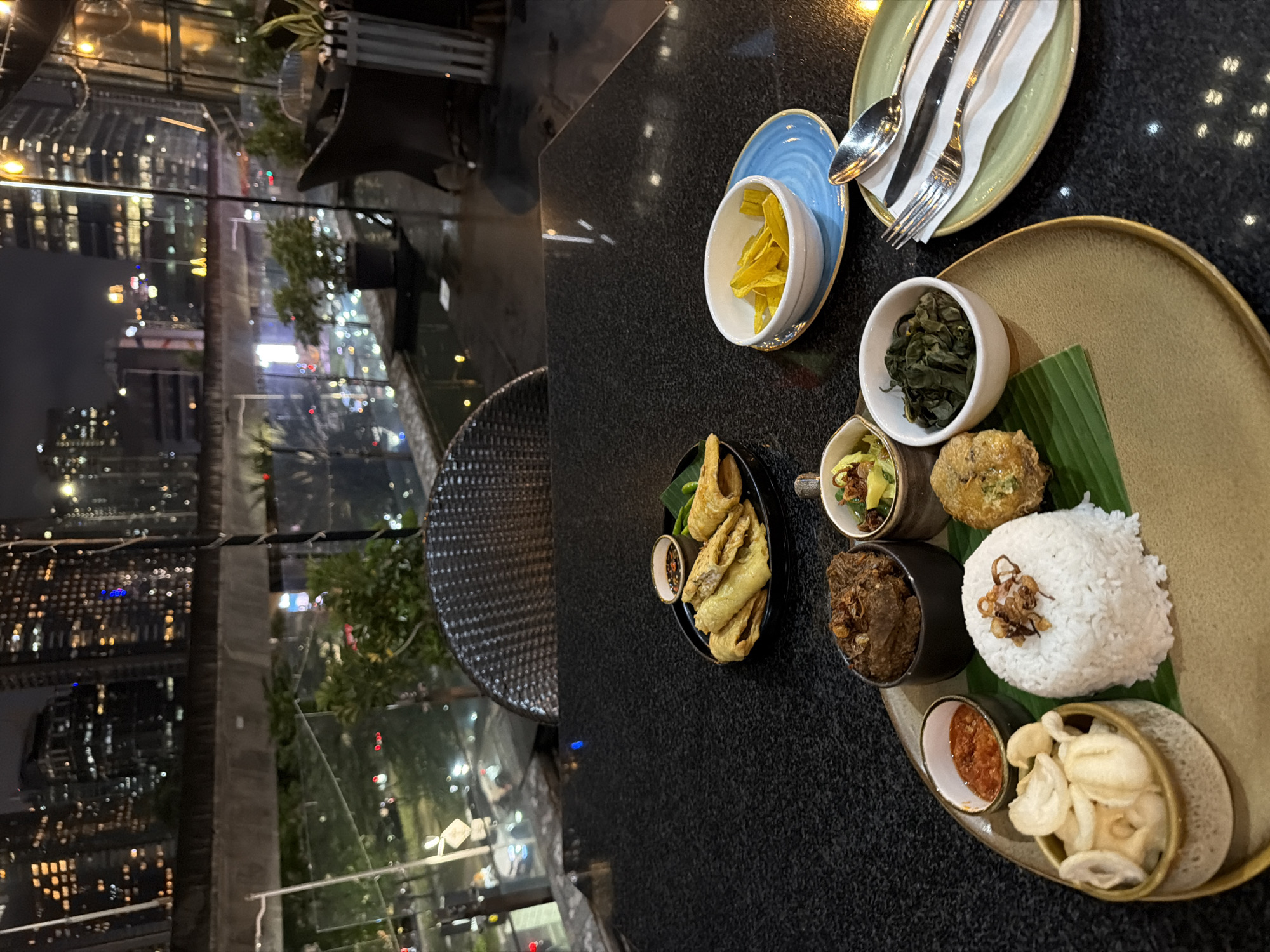 The photograph shows an overhead view of a meal presented on a black table in an indoor setting, with the night skyline of South Jakarta, Indonesia visible through a window. The meal consists of a plate with rice, side dishes served in small bowls, and crackers. Another smaller plate contains fried food with a dipping sauce. Utensils and a side dish of chips are also on the table. The environment suggests a restaurant or dining area with a view of the city.