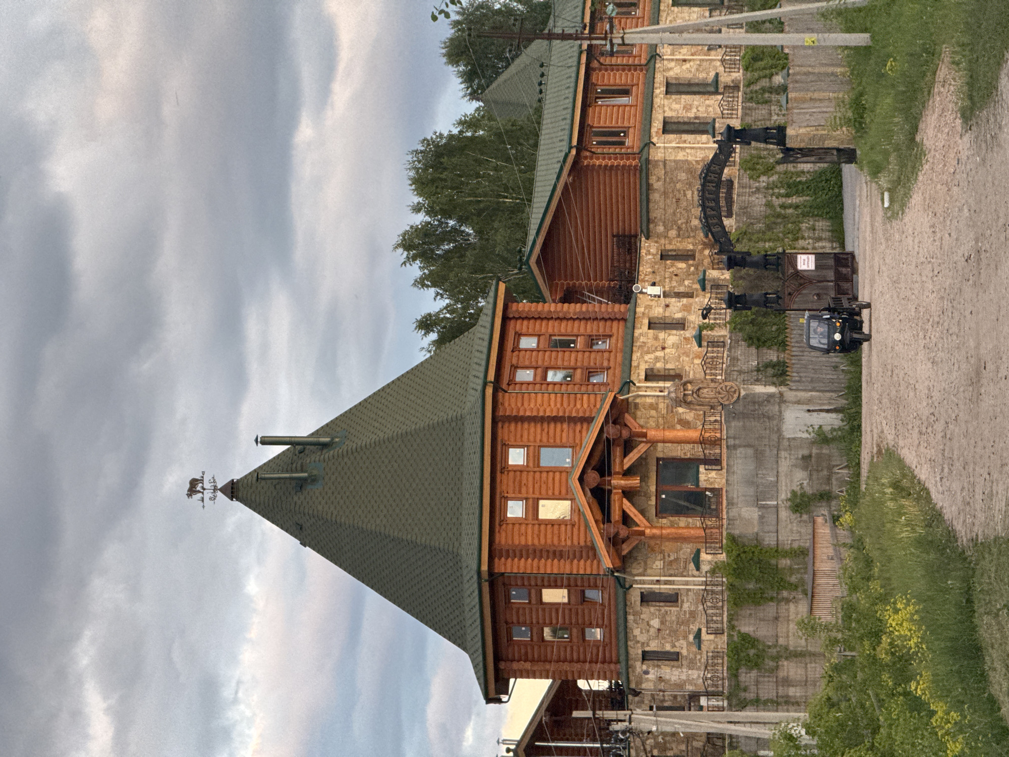 The image displays a rustic architectural complex, likely a lodge or large residence, situated in a rural setting under an overcast sky. The photo is rotated ninety degrees clockwise; when viewed upright, it shows the buildings from the perspective of a dirt road running alongside them.
The dominant structure on the left features a steep, dark green pyramidal roof, topped with a weather vane depicting a horse and rider. Several metal chimneys also rise from the roof. The upper stories are constructed with horizontal wooden logs or planks, characterized by numerous small, rectangular windows. The lower sections of the buildings are made of stone masonry, giving a robust and traditional appearance.
Towards the middle and right, the buildings continue with similar architectural elements, including stone bases, wooden upper sections, and balconies with intricate wrought-iron railings. A decorative wooden wagon wheel is mounted on a stone wall. Further right, a substantial black metal archway spans the dirt road, acting as an entrance or decorative element. The archway prominently displays the Russian word "БЕРЕГИТЕСЬ," meaning "BEWARE" or "TAKE CARE." Below the archway, a smaller, unreadable sign with red text is visible.
A small, black, three-wheeled enclosed utility vehicle is parked on the dirt road near the archway. A wooden utility pole stands beside the road. Green foliage, including trees in the background and grass and shrubs along the roadside, indicates a natural environment. The sky is uniformly gray and overcast, suggesting diffused light, possibly during morning or late afternoon. No people are visible, and the scene appears quiet and still. The overall impression is one of a well-maintained, substantial property with a strong, rustic aesthetic, typical of a rural Russian setting like Kamysh.