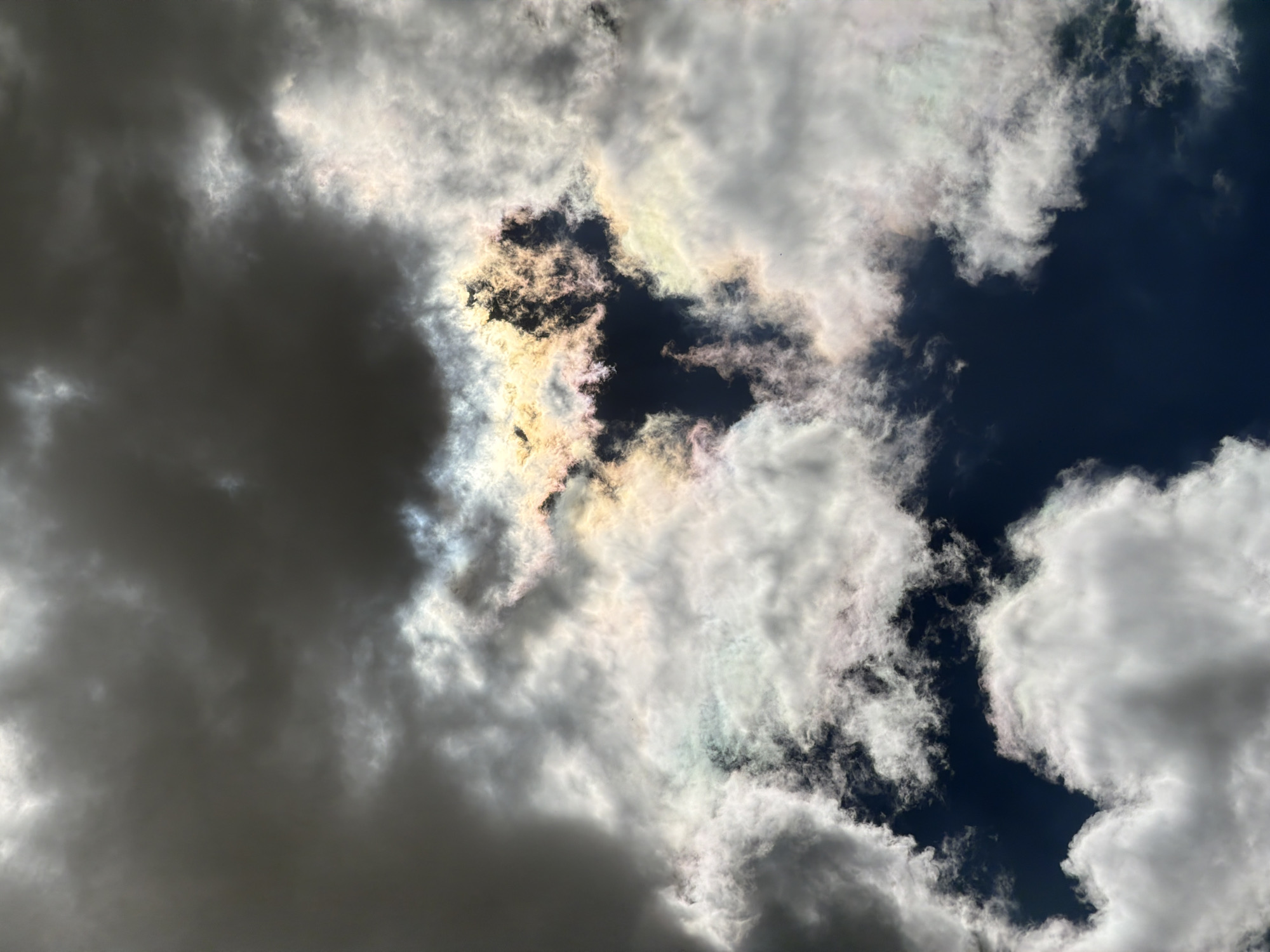 The image captures a dramatic sky scene with a mixture of dark storm clouds and bright, iridescent clouds. In the center, sunlight pierces through a gap in the clouds, creating a vibrant display of rainbow-like colors within the cloud formations. The dark clouds on the upper left and surrounding areas suggest an approaching or receding storm. The overall impression is one of dynamic atmospheric conditions. The image does not contain any people, subjects, or objects that would indicate a specific location within Burgas, Bulgaria. It is a natural sky phenomenon.