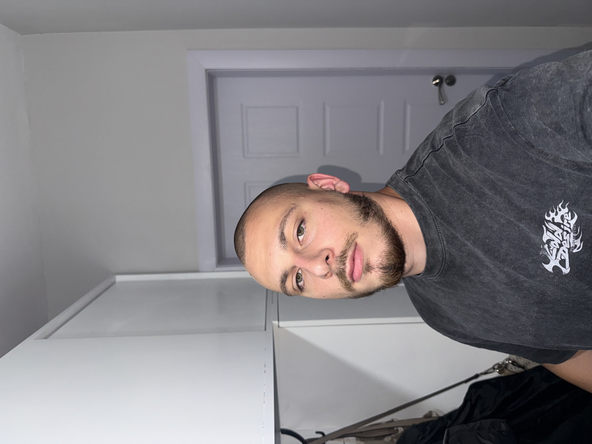 A man with a shaved head, a goatee, and a mustache stands in front of a door. He is wearing a dark grey t-shirt with a graphic design. The room appears to be a closet or a hallway, with white cabinets on the left and a closed door on the right. The lighting is dim, casting shadows on the man's face. The image does not provide any information about the location in Stara Zagora, Bulgaria.