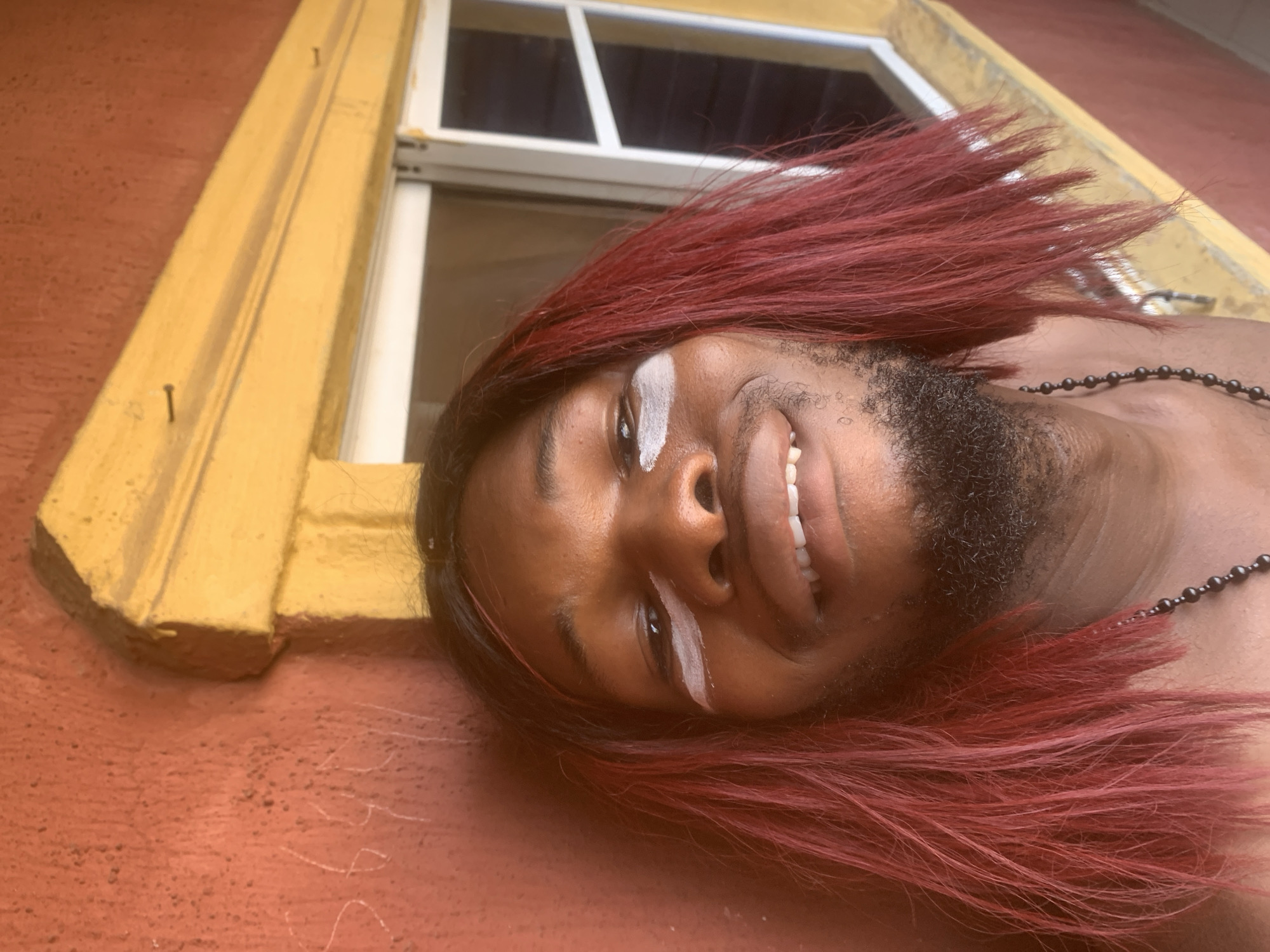 The image presents a tilted, close-up portrait of a person with dark skin, likely male, against a reddish-orange wall in Nkpor, Nigeria. The individual is smiling broadly, showing white teeth, and appears cheerful. They have long, vibrant red hair or a wig that flows across the background, and white paint or makeup streaks are visible over and below the left eyebrow, and above the right. Facial hair, appearing as a beard or stubble, is also present. A dark, beaded necklace is worn around the neck.
The background features part of a building with a bright yellow window trim, from which several nails protrude. Inside the yellow trim, a white window frame is visible, with a dark interior seen through the glass. The wall itself has a textured, reddish-orange finish. Faint white writing, possibly "1995 S," is discernible on the wall in the lower right section of the image. The scene is brightly lit, indicating daytime. The composition suggests an artistic or expressive pose.